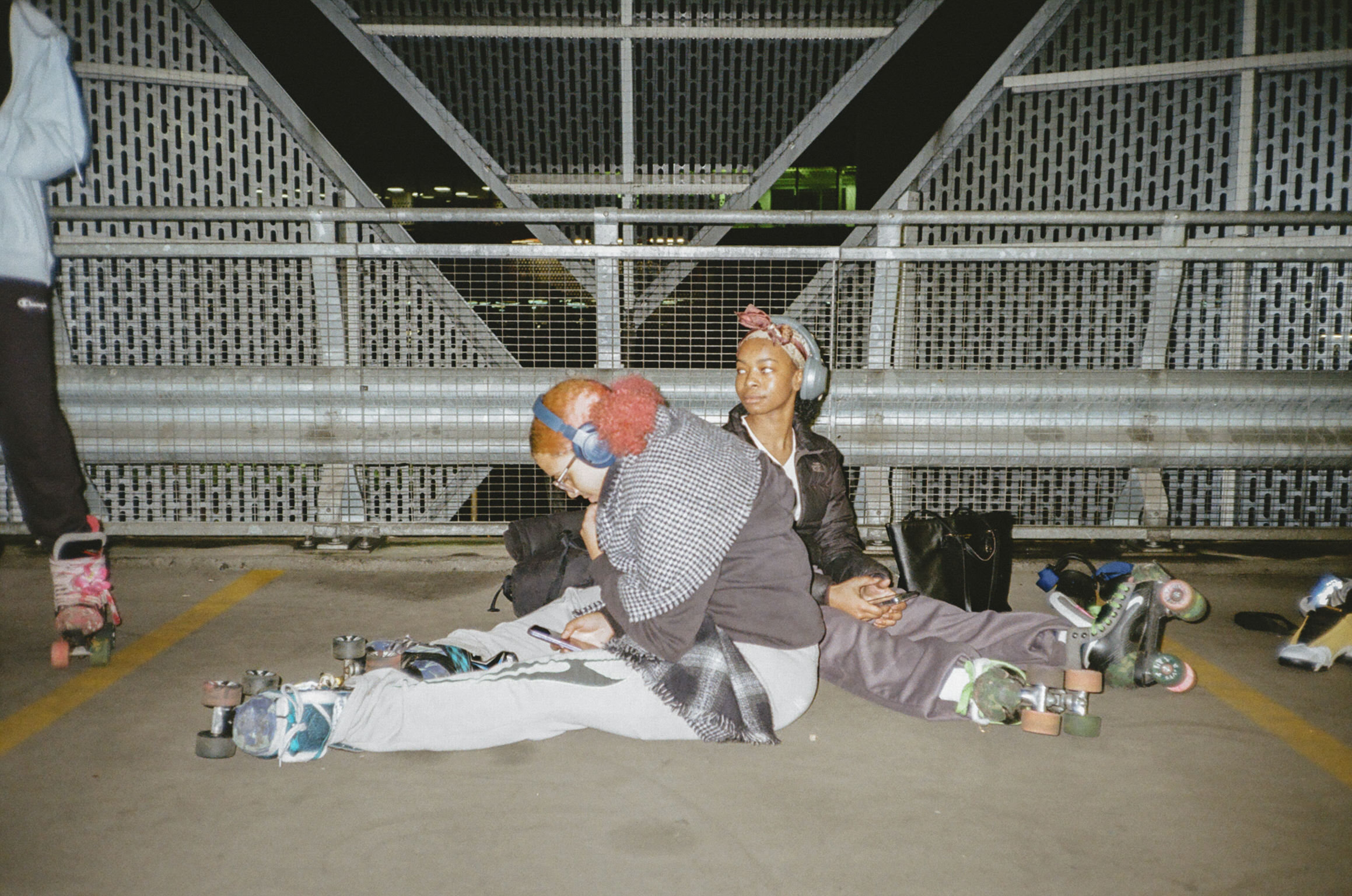 Two people sitting on concrete floor under metal bridge structure with belongings scattered around them.