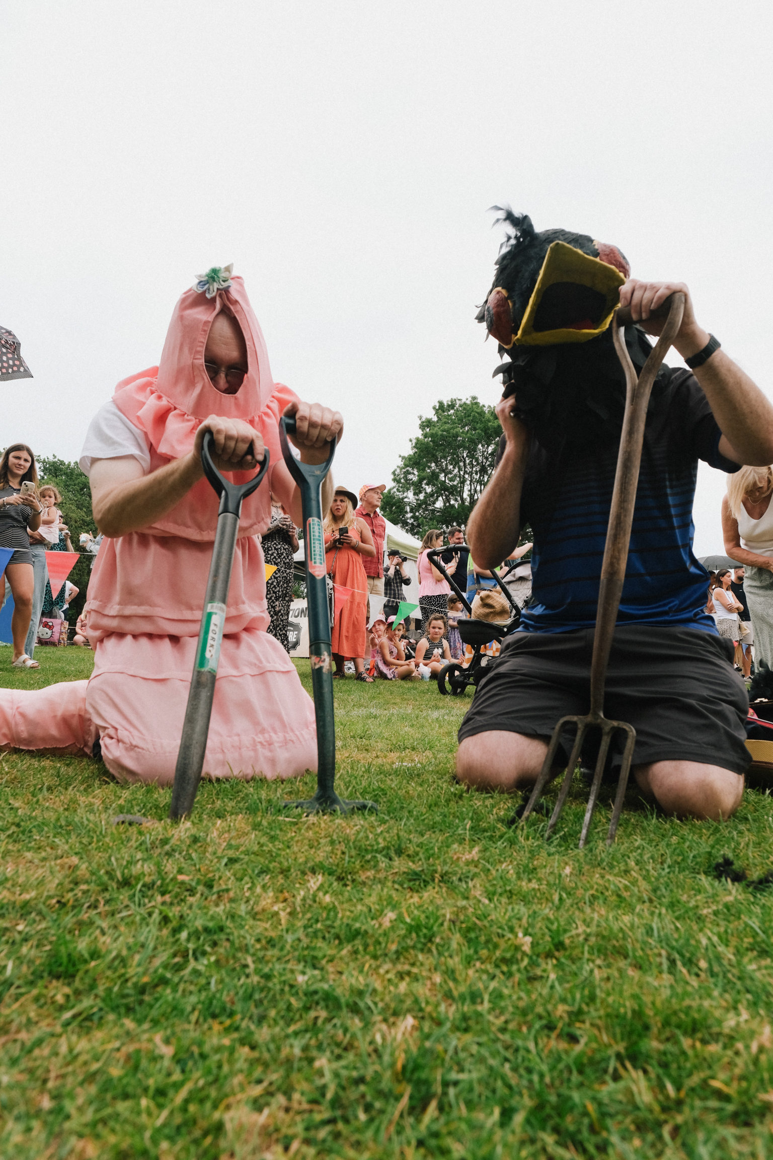 Two people in medieval costume kneeling on grass, holding swords. Person on left wears pink dress, person on right wears blue tunic and helmet.
