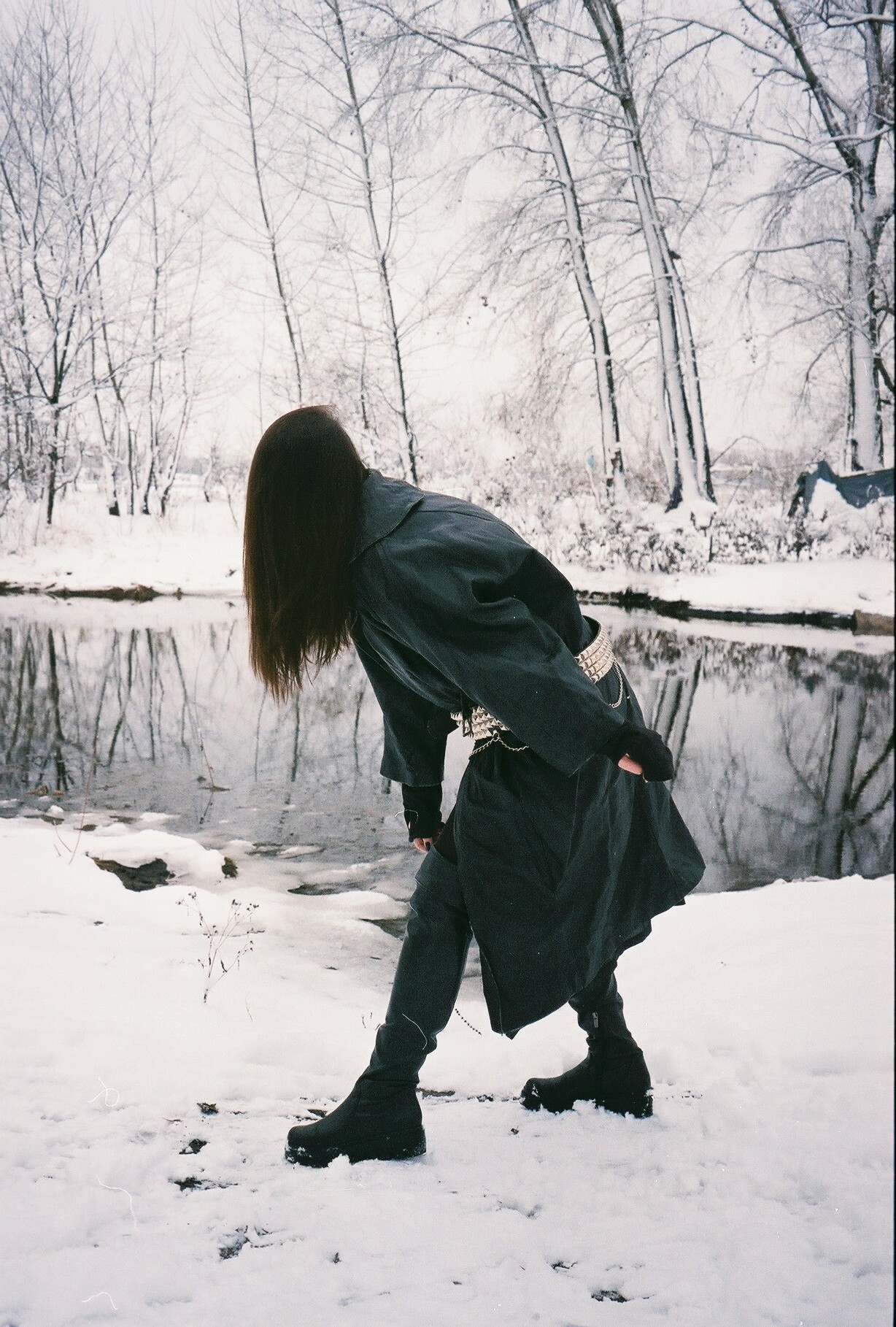 Snowy forest with bare trees, dark figure standing in foreground wearing hooded black coat, standing in snow by a pond.