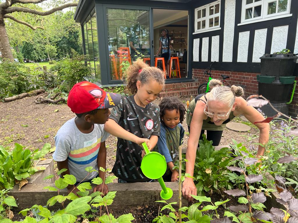 Adult and three children tending to plants in raised garden bed, using green watering can. Tudor-style house with conservatory in background.