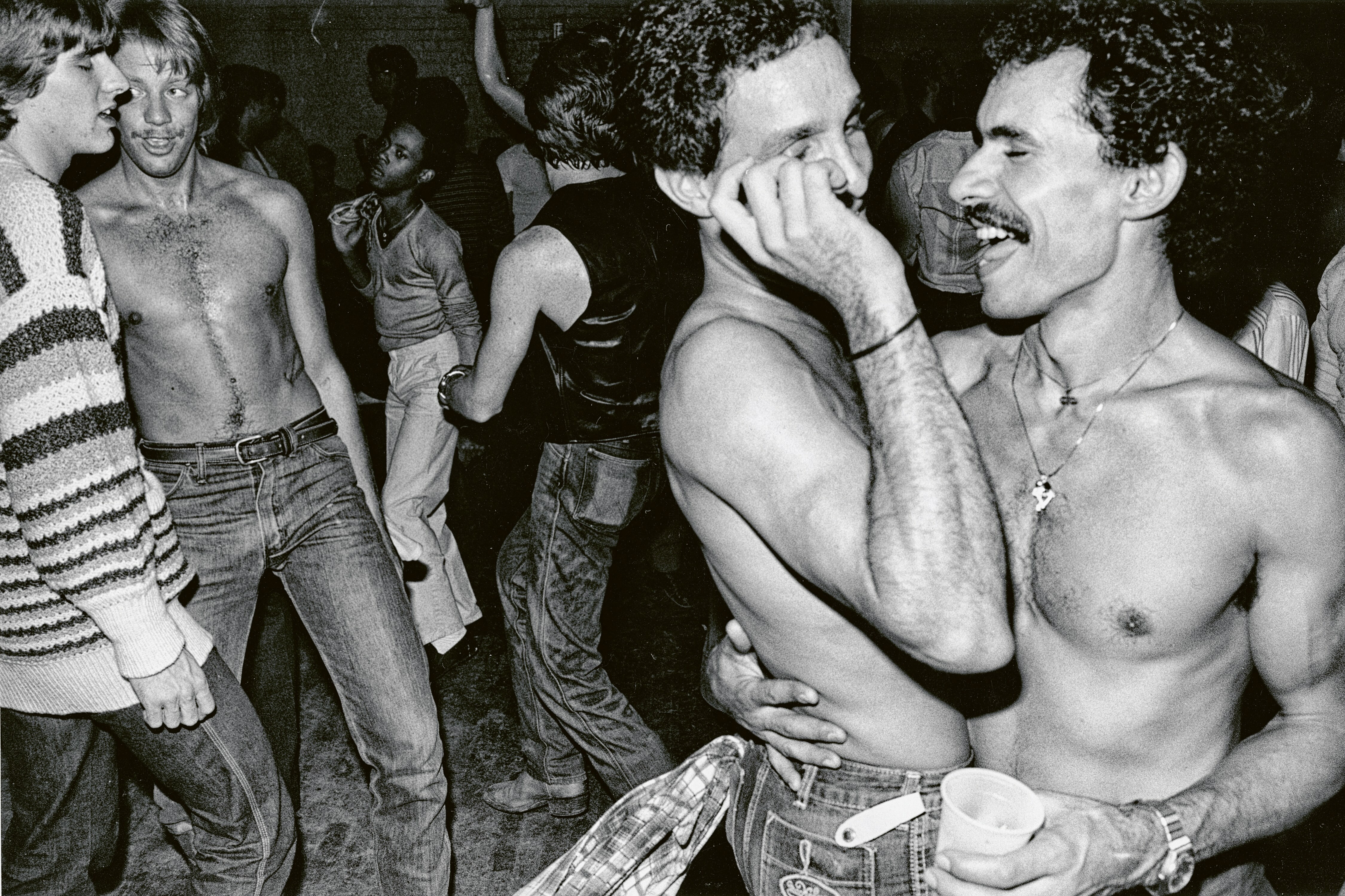 Black and white image showing a group of shirtless men socialising, some laughing.