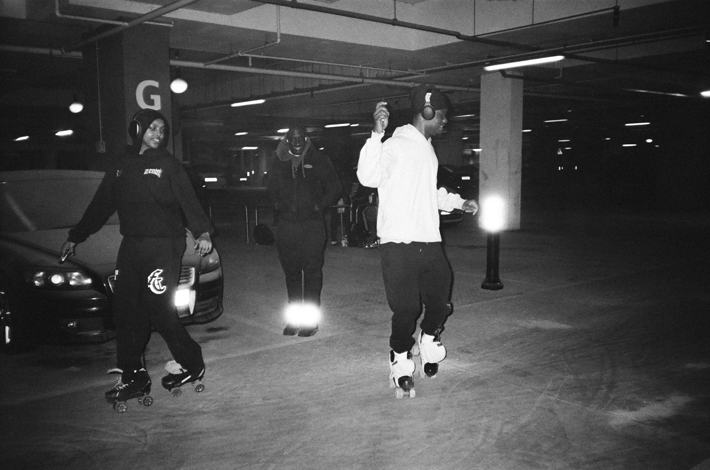 Black and white image of two people roller skating in an indoor car park with concrete pillars and fluorescent lighting overhead.