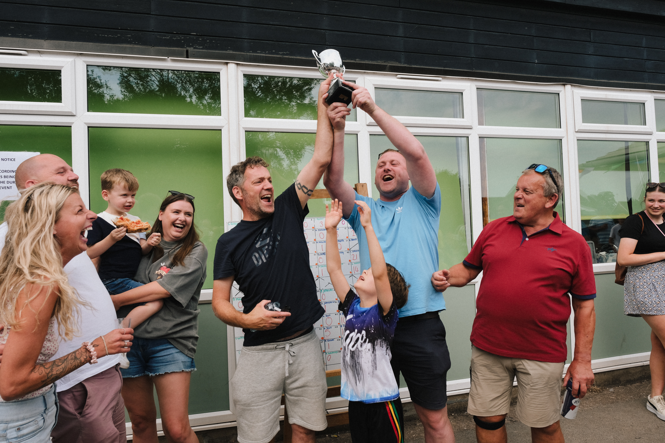 Man in light blue shirt holds trophy aloft whilst group of people celebrate around him outside building with large windows.
