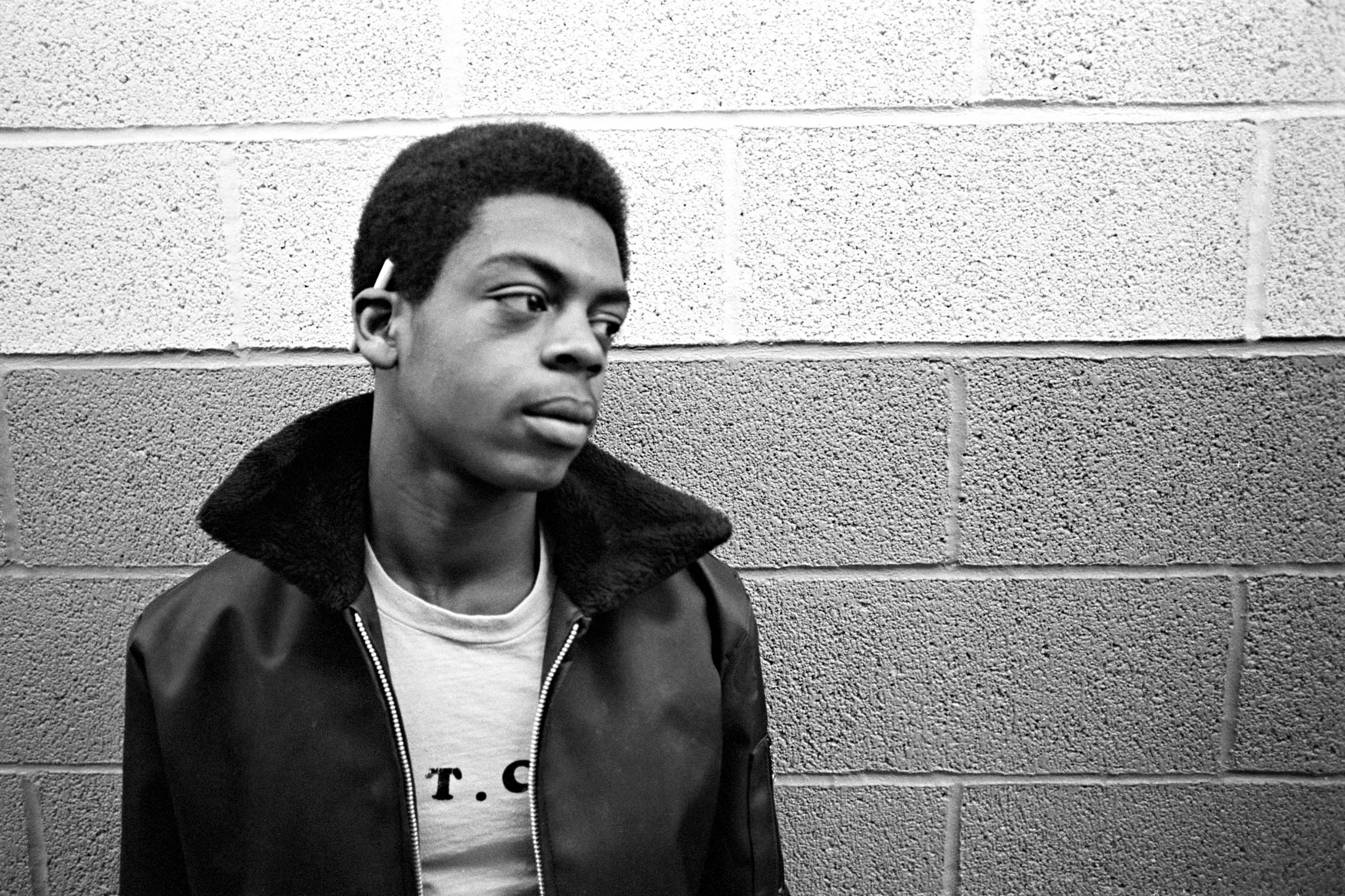 Black and white portrait of young Black man in hooded jacket against brick wall, looking to the side with earbuds in.