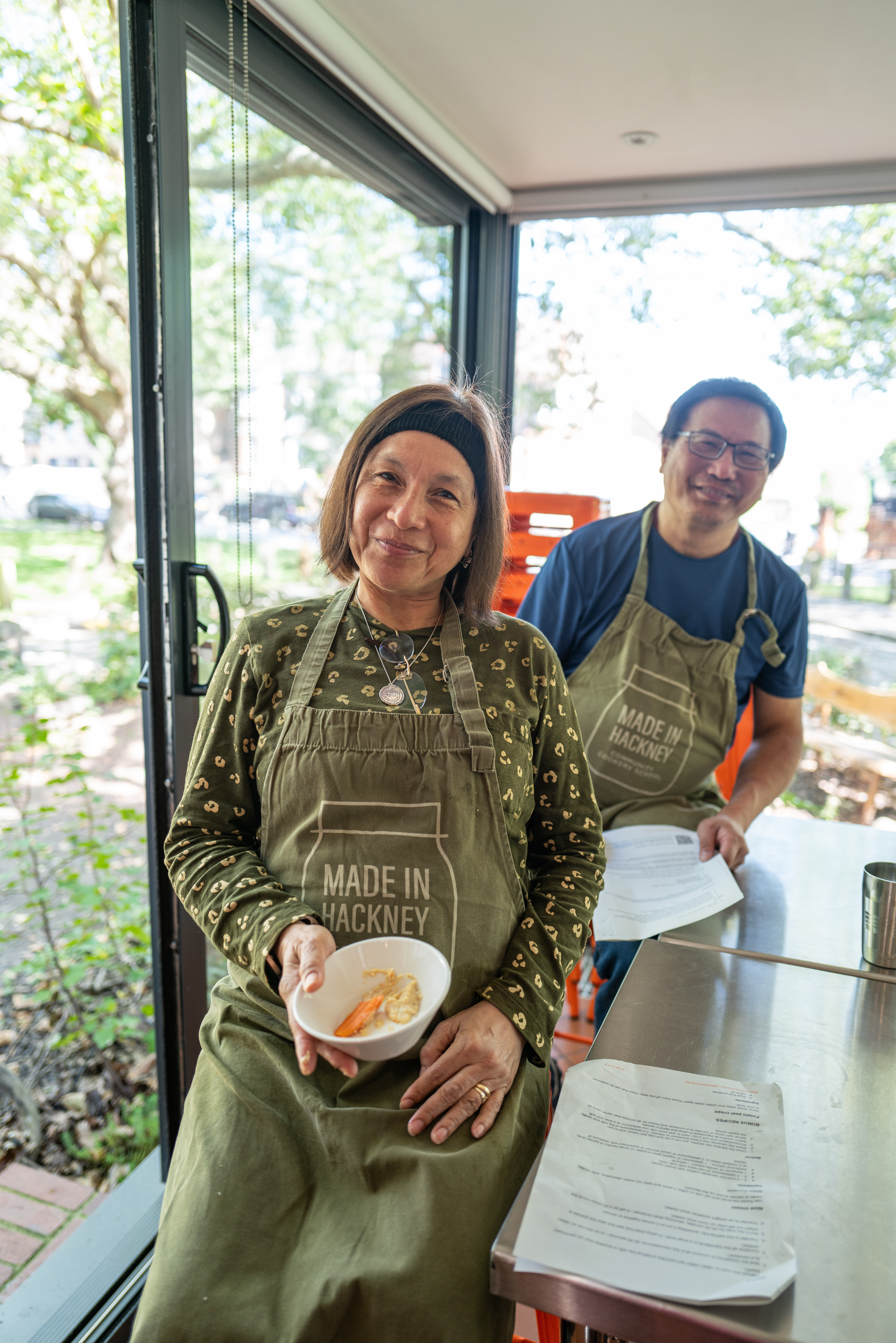 Woman and man wearing green "Made in Hackney" aprons in bright kitchen with large windows overlooking leafy street.