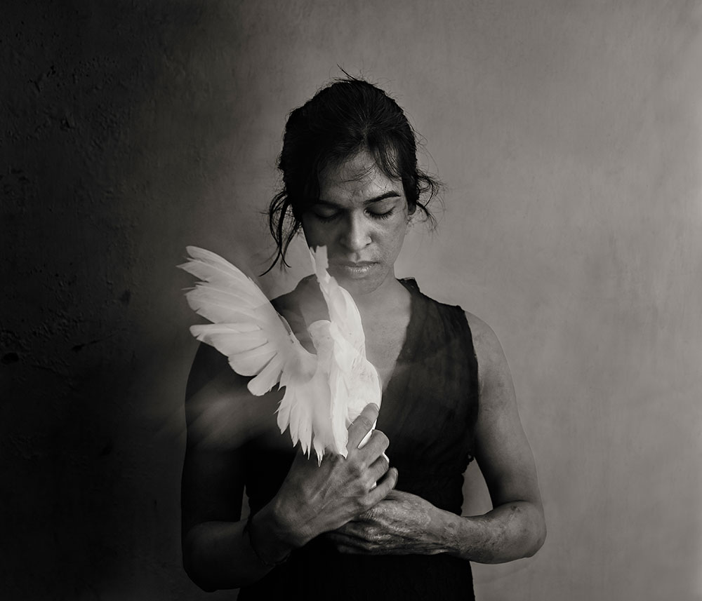 Woman in black top holding white dove against muted grey-green background, soft lighting creates atmospheric mood.