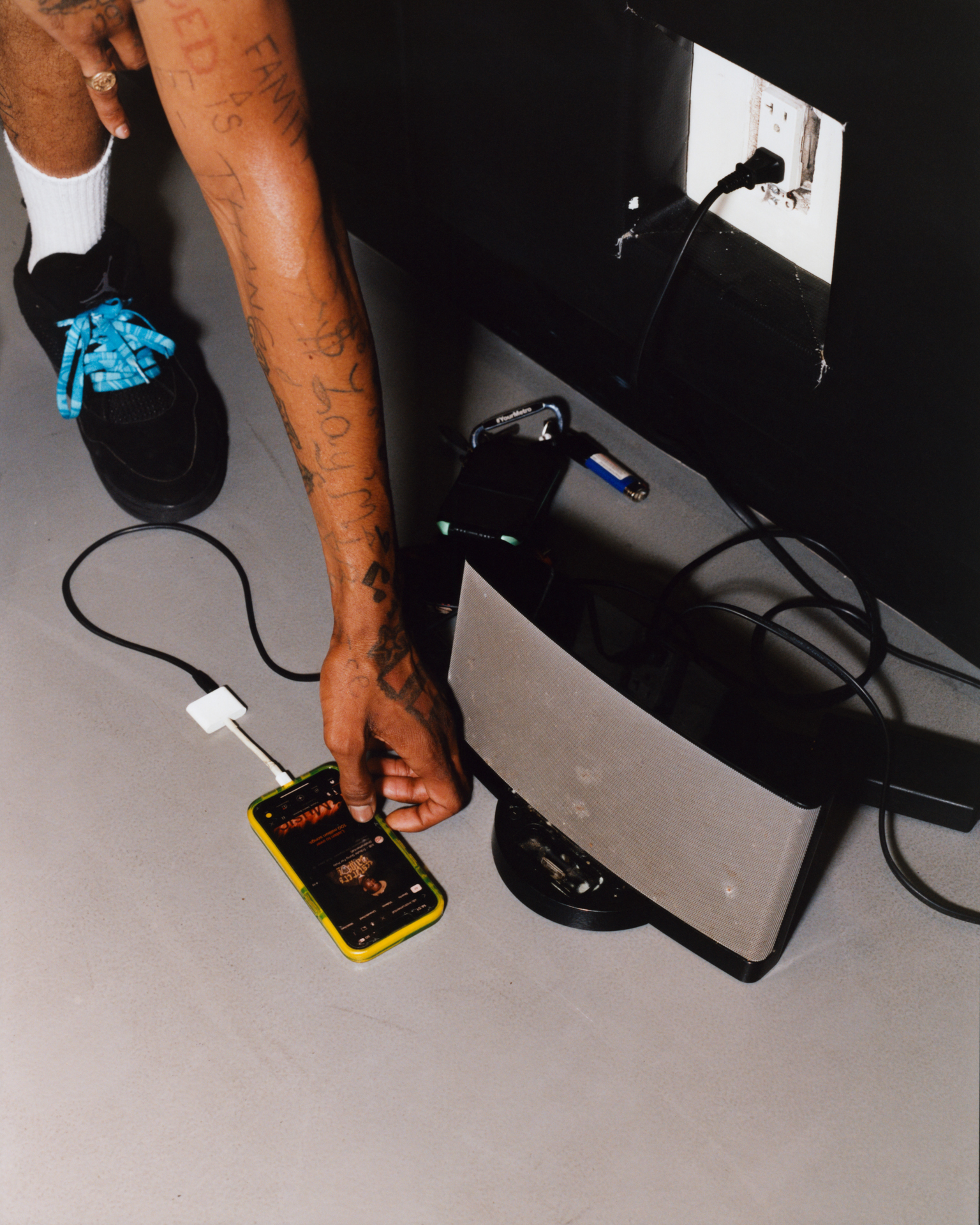 Person's tattooed arm reaching towards yellow mobile phone on white desk with black laptop, cables, and gaming controller visible.