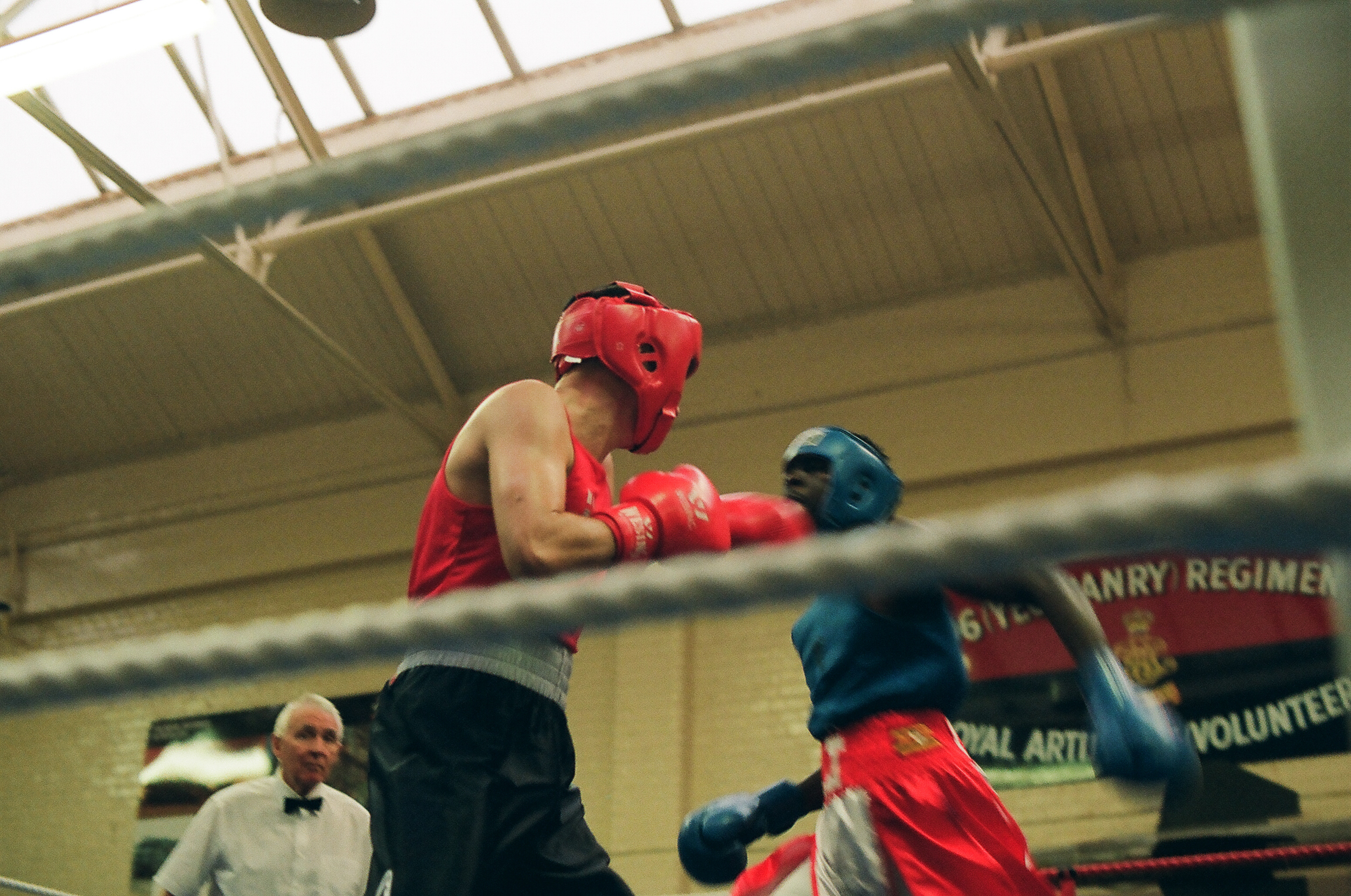 Two boxers sparring in ring, one wearing red headgear and gloves, other in blue. Referee watches from ringside in indoor venue.