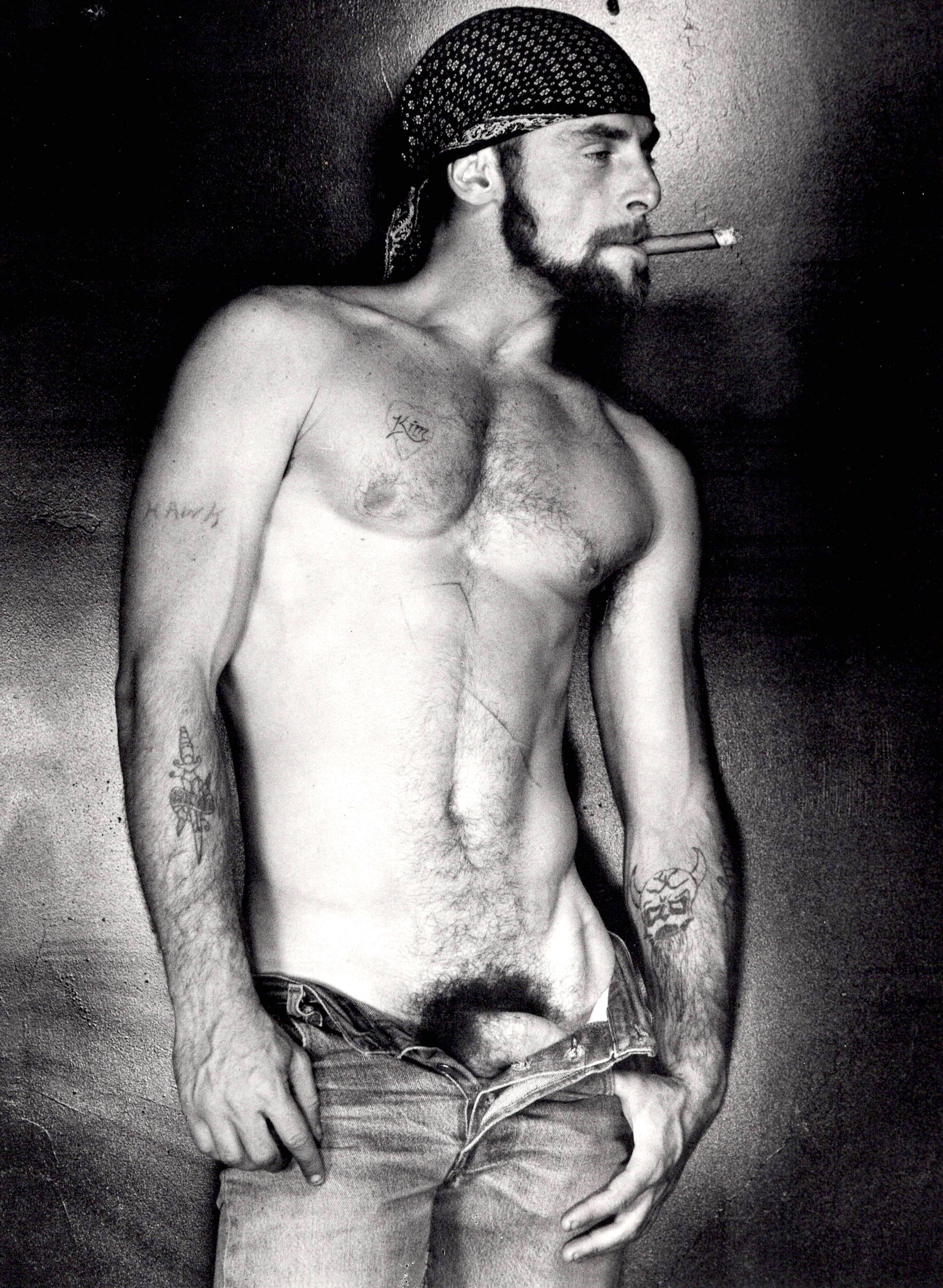 Black and white portrait of shirtless man with beard, bandana, and cigarette, wearing unbuttoned jeans, looking to the side.