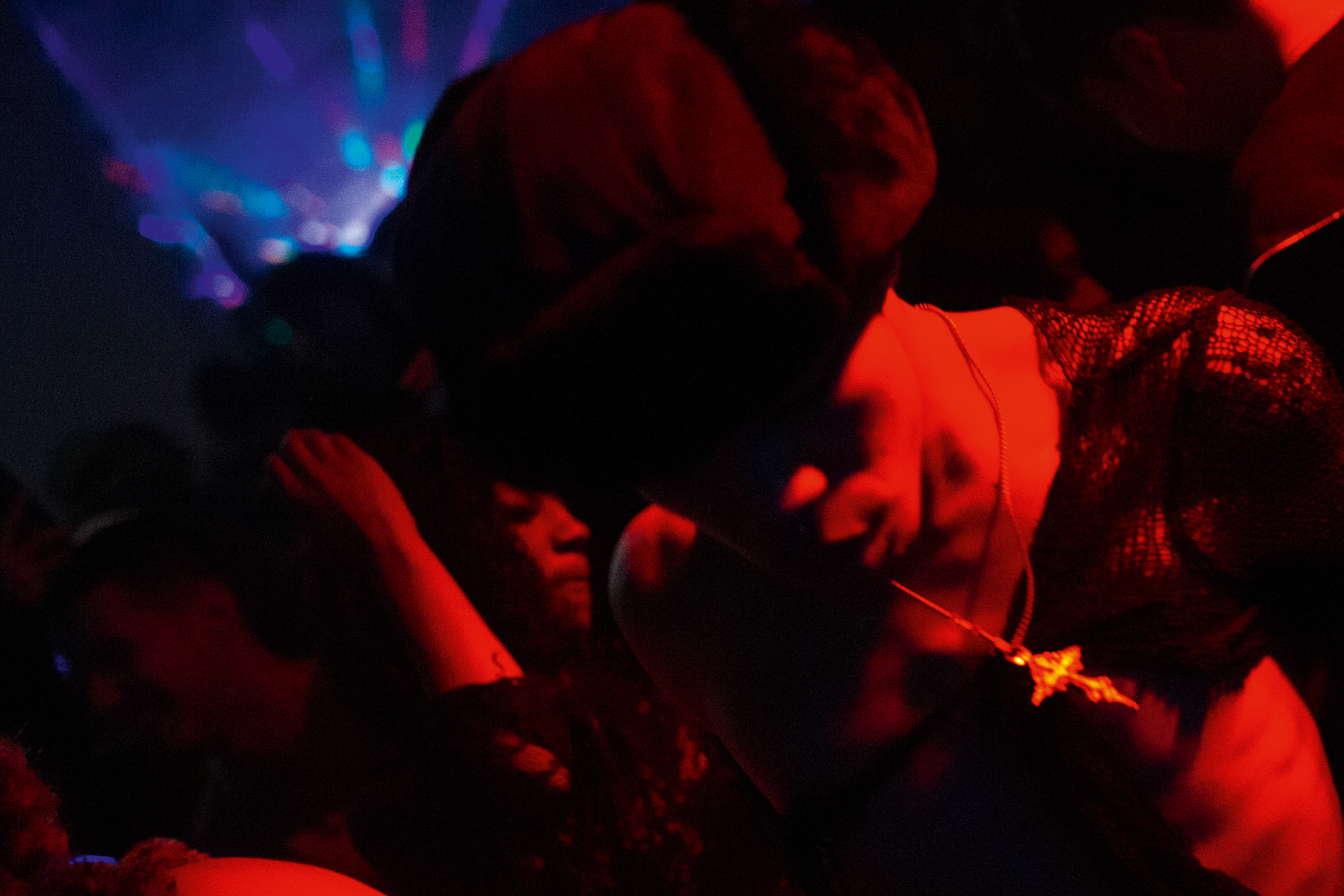Crowded nightclub scene with intense red lighting illuminating people dancing, blue lights visible in background, dark shadows throughout.