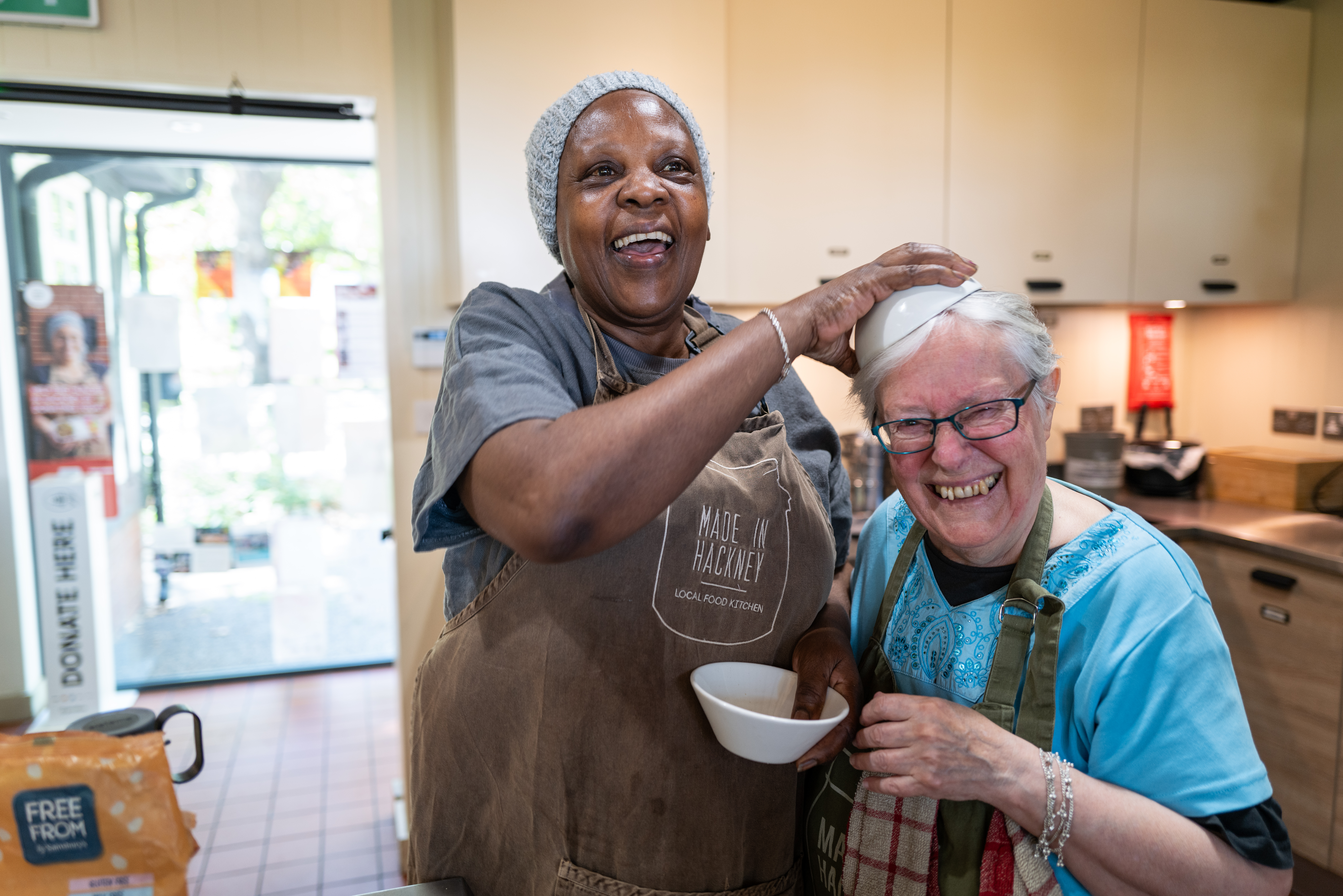 Black woman in hairnet and apron serves soup to smiling white woman in blue top at community kitchen serving counter.