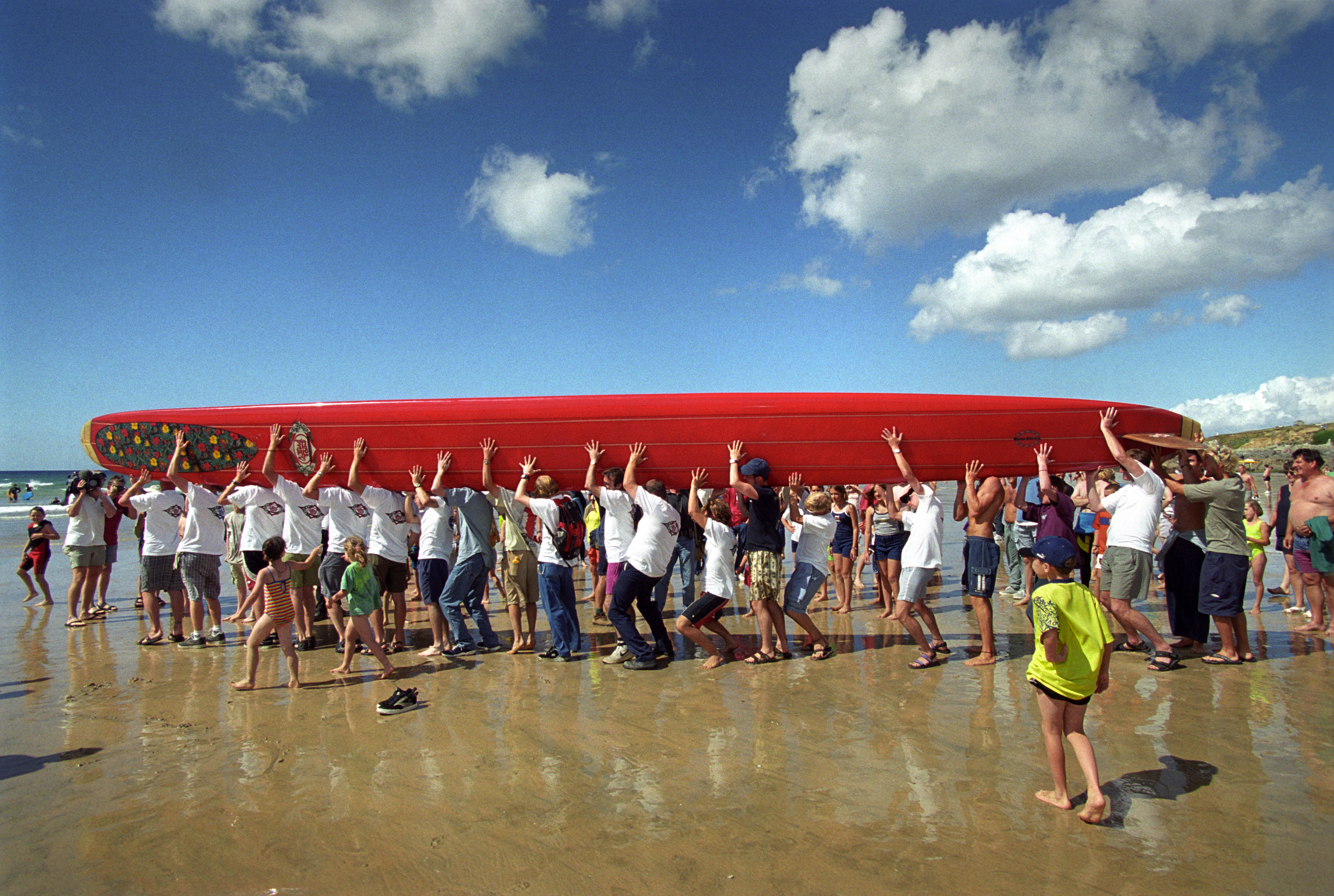 Crowd of people carrying large red surfboard on beach under cloudy sky.