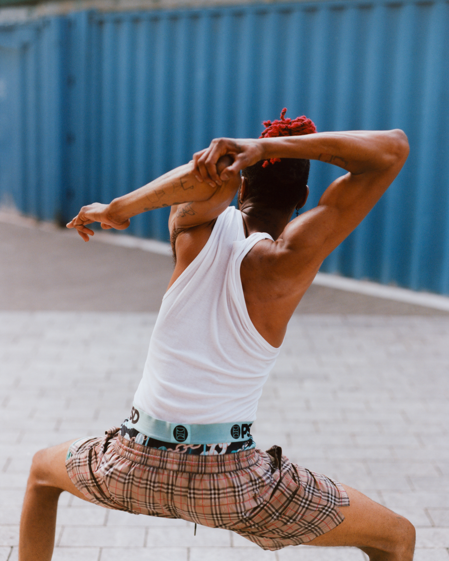 Person in white tank top and plaid shorts stretching arms behind head, blue corrugated fence in background.