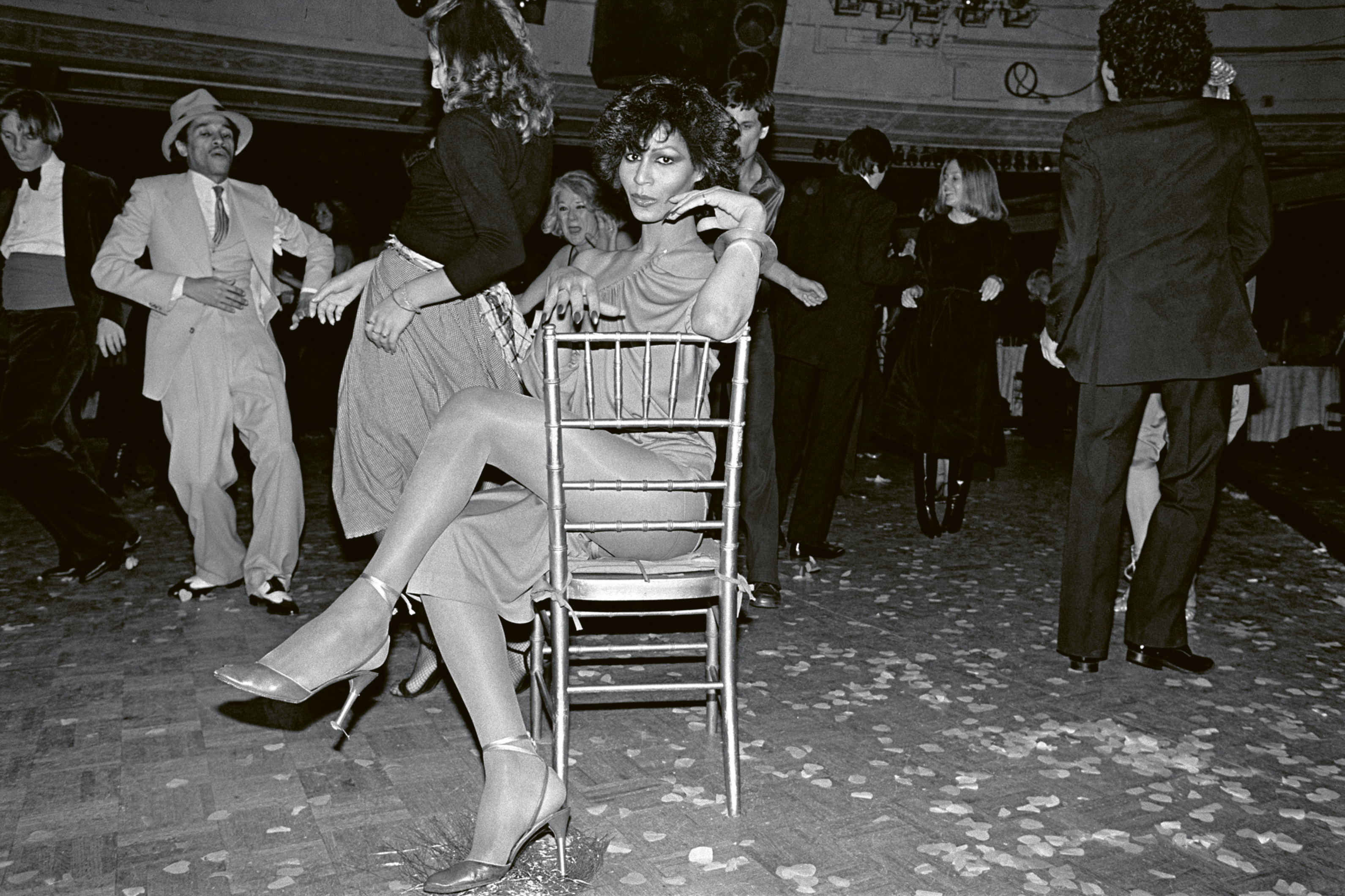 Black and white image of a woman sitting on a chair in an indoor setting, surrounded by people.
