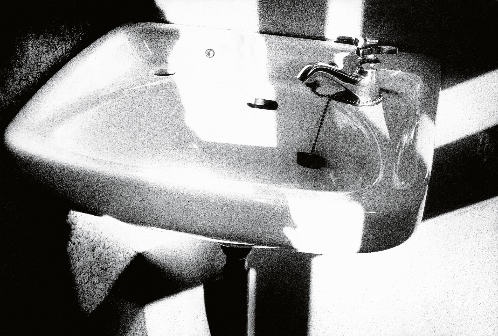 Black and white image of bathroom sink with chrome tap, mounted on pedestal base, strong shadows cast across white surfaces.