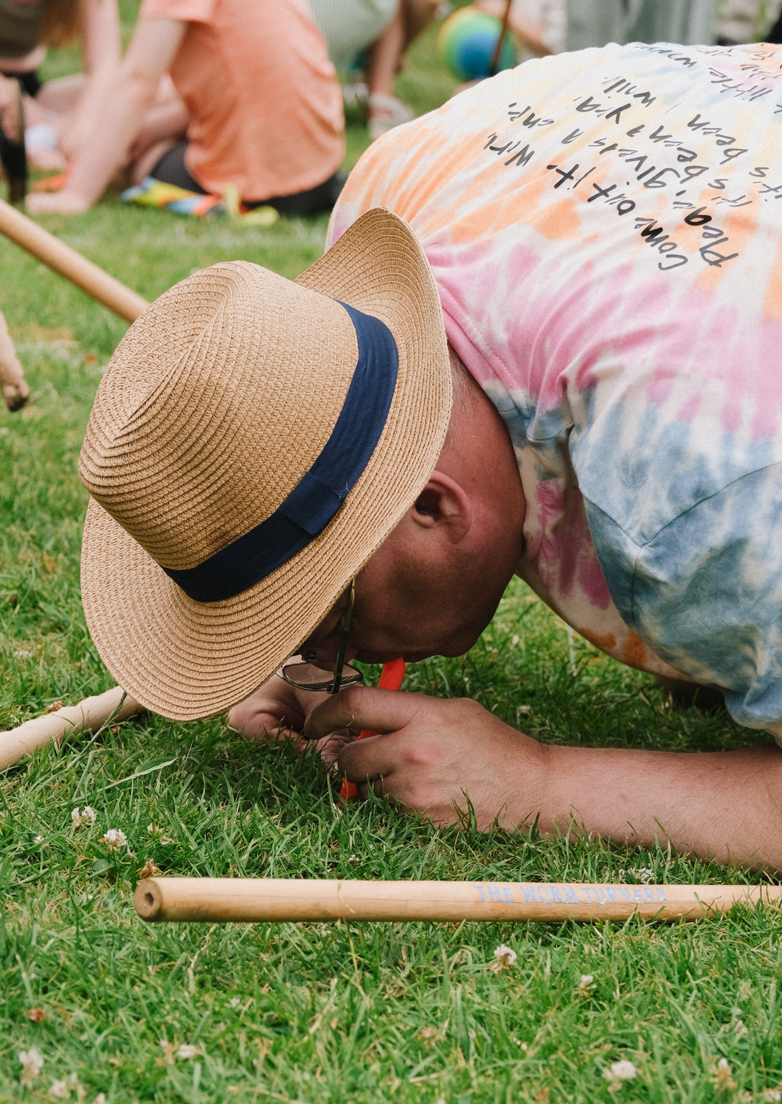 Person in straw hat with navy band crawling under limbo stick on grass, wearing tie-dye shirt with handwritten text.