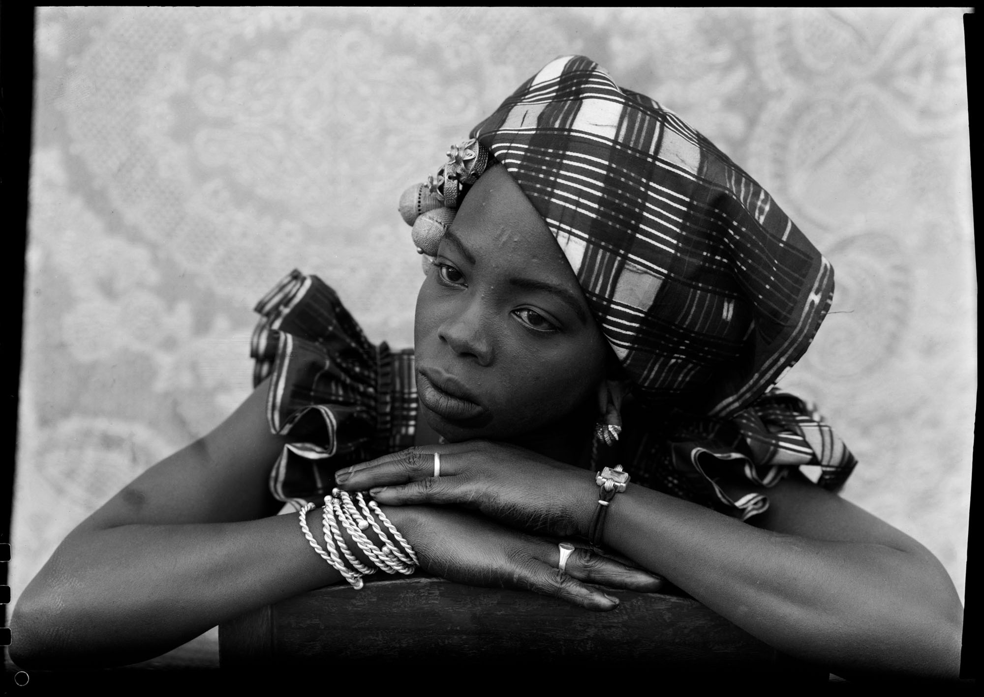 Black and white portrait of woman wearing patterned headwrap and jewellery, arms crossed, resting chin on hands.