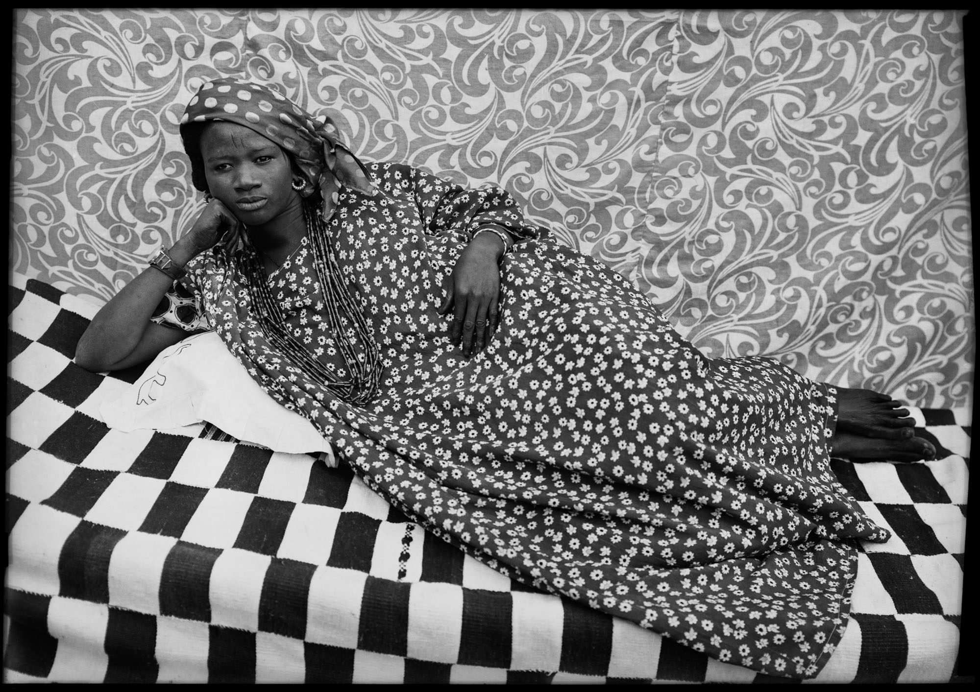 Black and white image: woman in polka dot dress reclining on chequered surface against ornate floral wallpaper background.