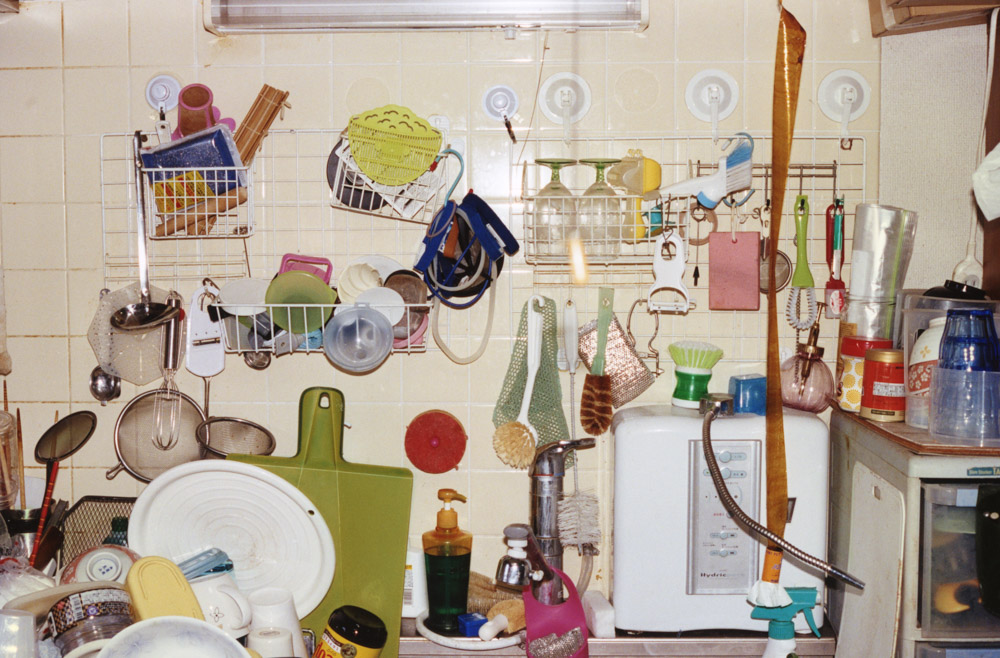Kitchen with white tiled walls covered in wall-mounted wire baskets, hooks, and shelves holding colourful utensils and cookware.
