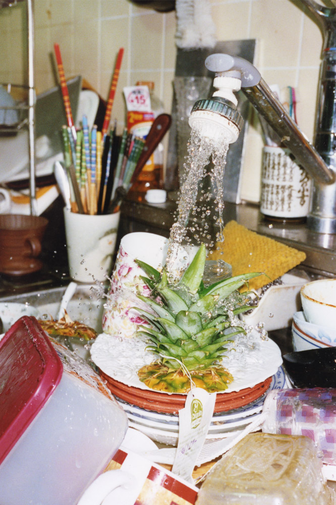 Water spraying from kitchen tap onto pineapple top in white bowl, paintbrushes in pot, cluttered kitchen counter with dishes.