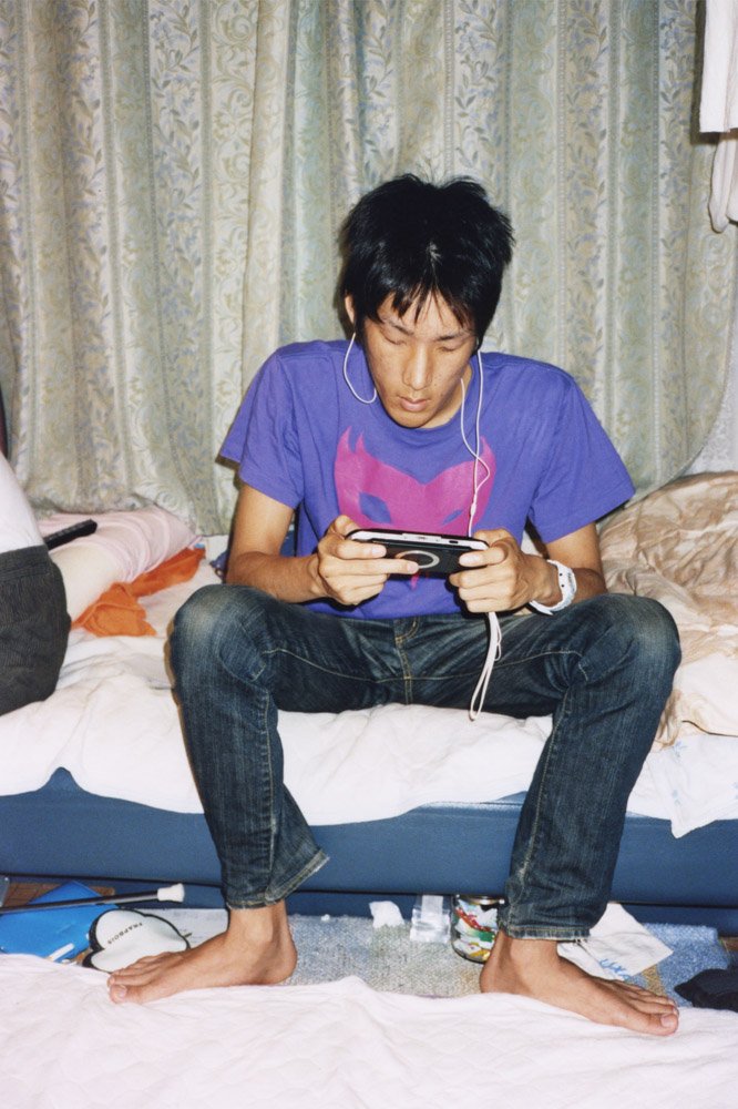 Man in purple t-shirt and jeans sits on bed playing handheld gaming device with white earphones, patterned curtains behind.