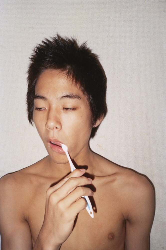 Young Asian man with short dark hair holding white toothbrush to mouth, eyes closed, bare shoulders visible against pale background.