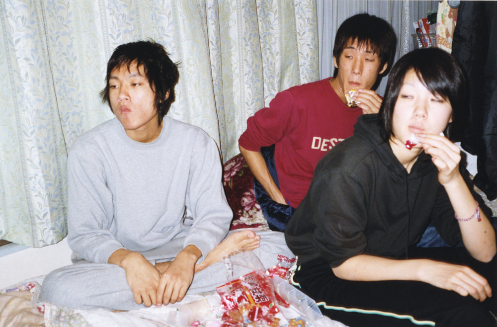 Three young people sitting on bed with floral bedding. Left person in grey jumper, middle in red top, right in black shirt eating.
