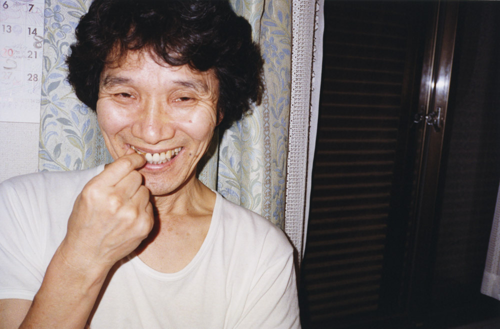 Smiling person with dark curly hair wearing white top, hand near chin, calendar visible on wall behind them.