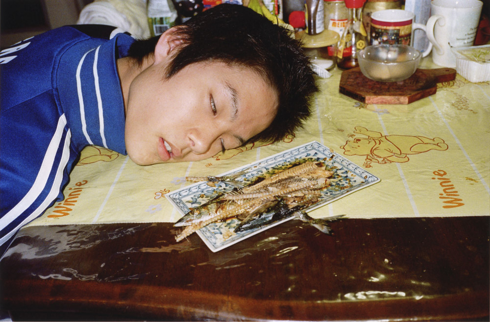 Person in blue tracksuit sleeping at table with head resting on yellow tablecloth, jigsaw puzzle pieces scattered nearby.