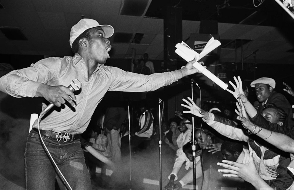 Performer in white cap and shirt leans forward on stage, reaching towards crowd with outstretched arms in black and white image.