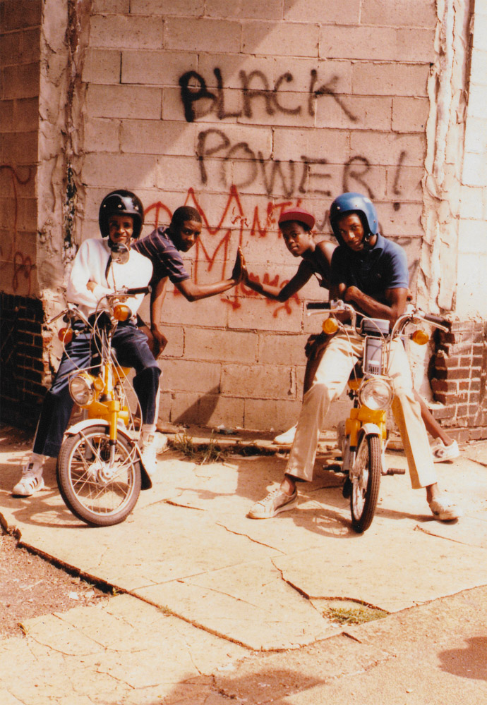 Four people on yellow mopeds giving high-fives in front of brick wall with black and red graffiti text.