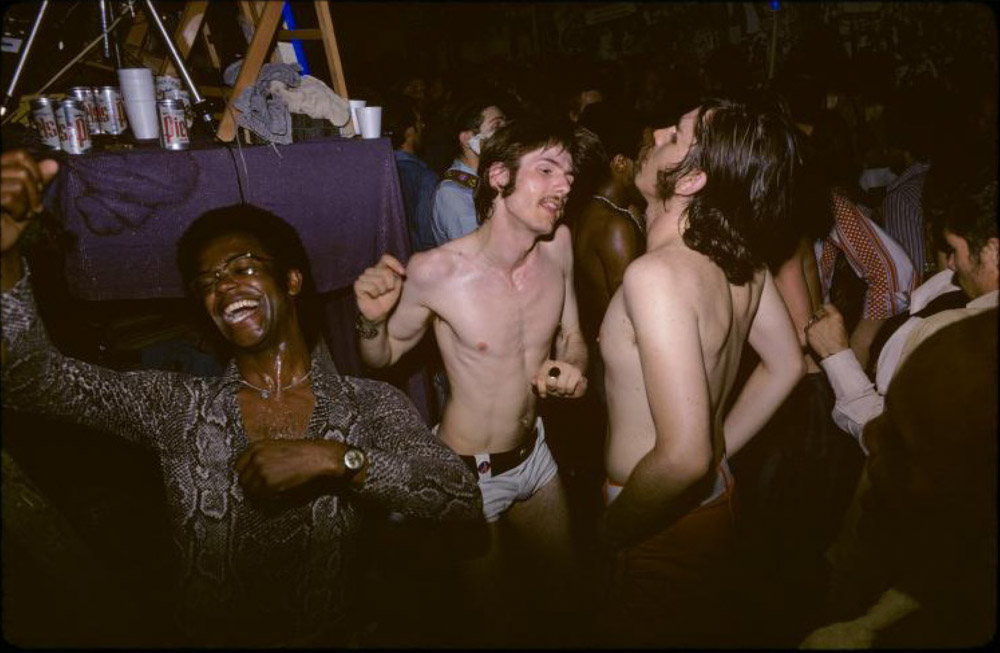 Group of people dancing at crowded indoor party, some shirtless, dim lighting with purple fabric visible in background.