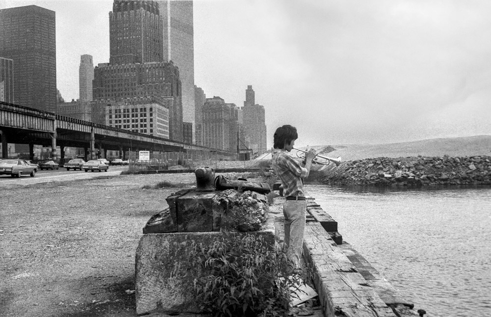 Black and white image showing person sitting on concrete pier with city skyline and bridge in background across water.