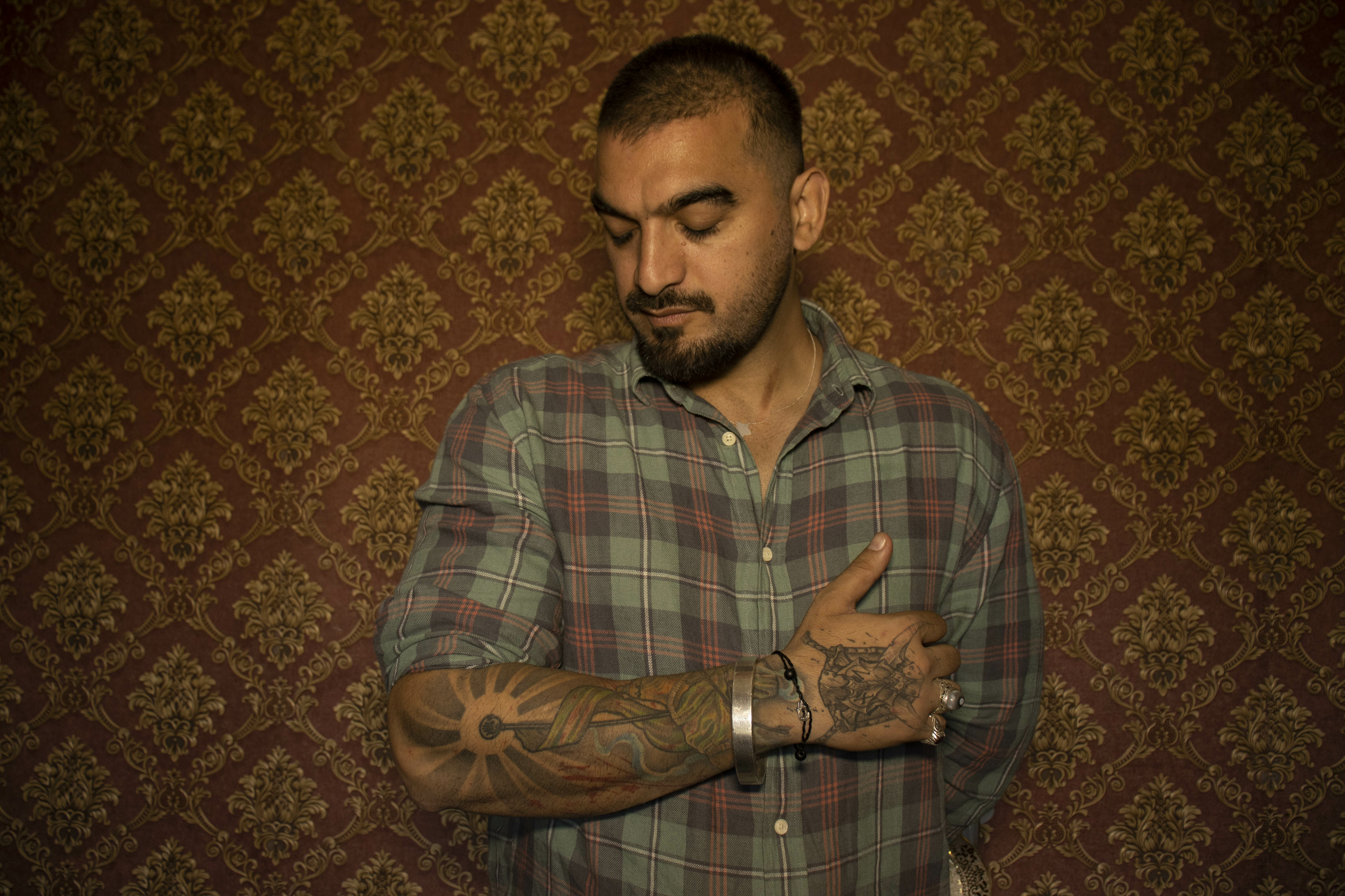 A bearded man in a checked shirt with tattoos on his arms, standing in front of a patterned background.