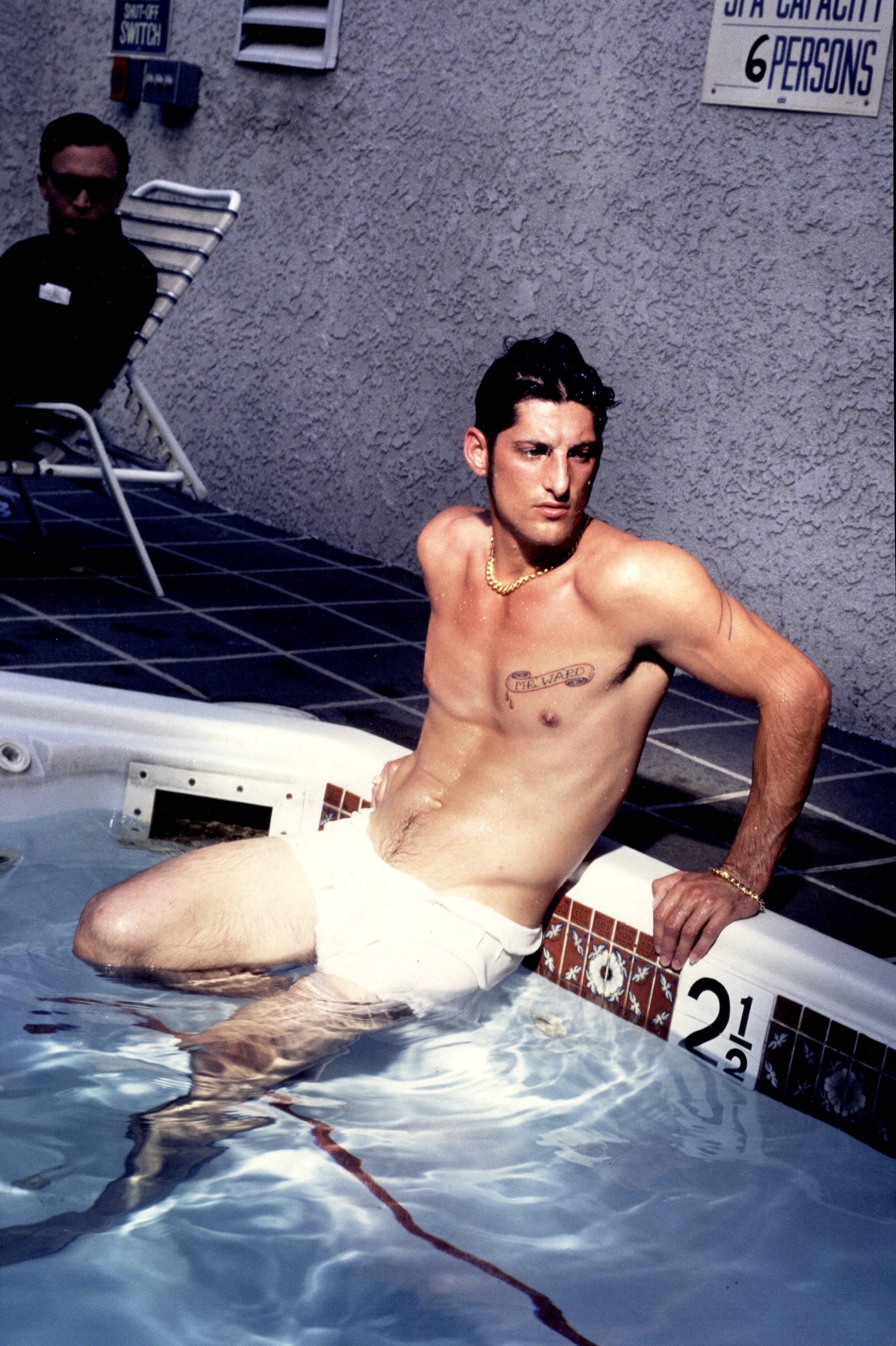 Shirtless man sitting on edge of swimming pool with legs in water, another person in background on deck chair