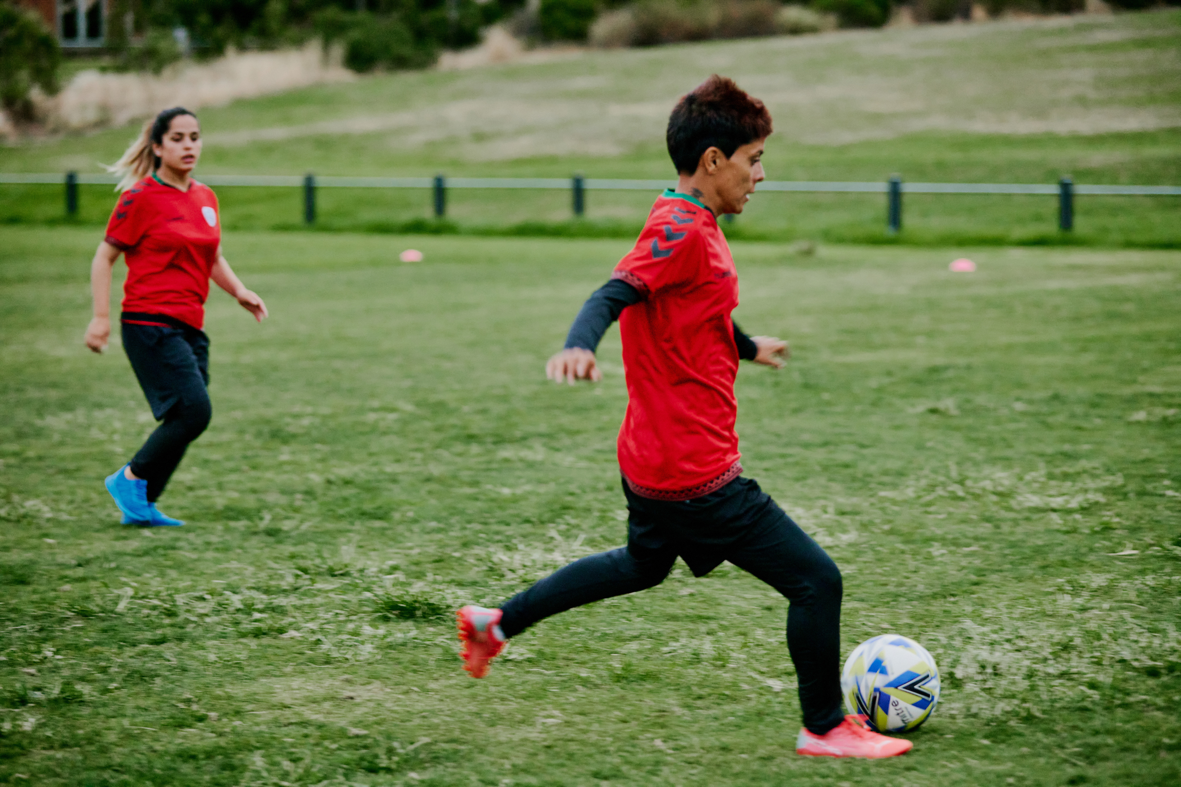 Two players in red shirts and dark trousers running on grass field, one dribbling football, with fence and buildings in background.