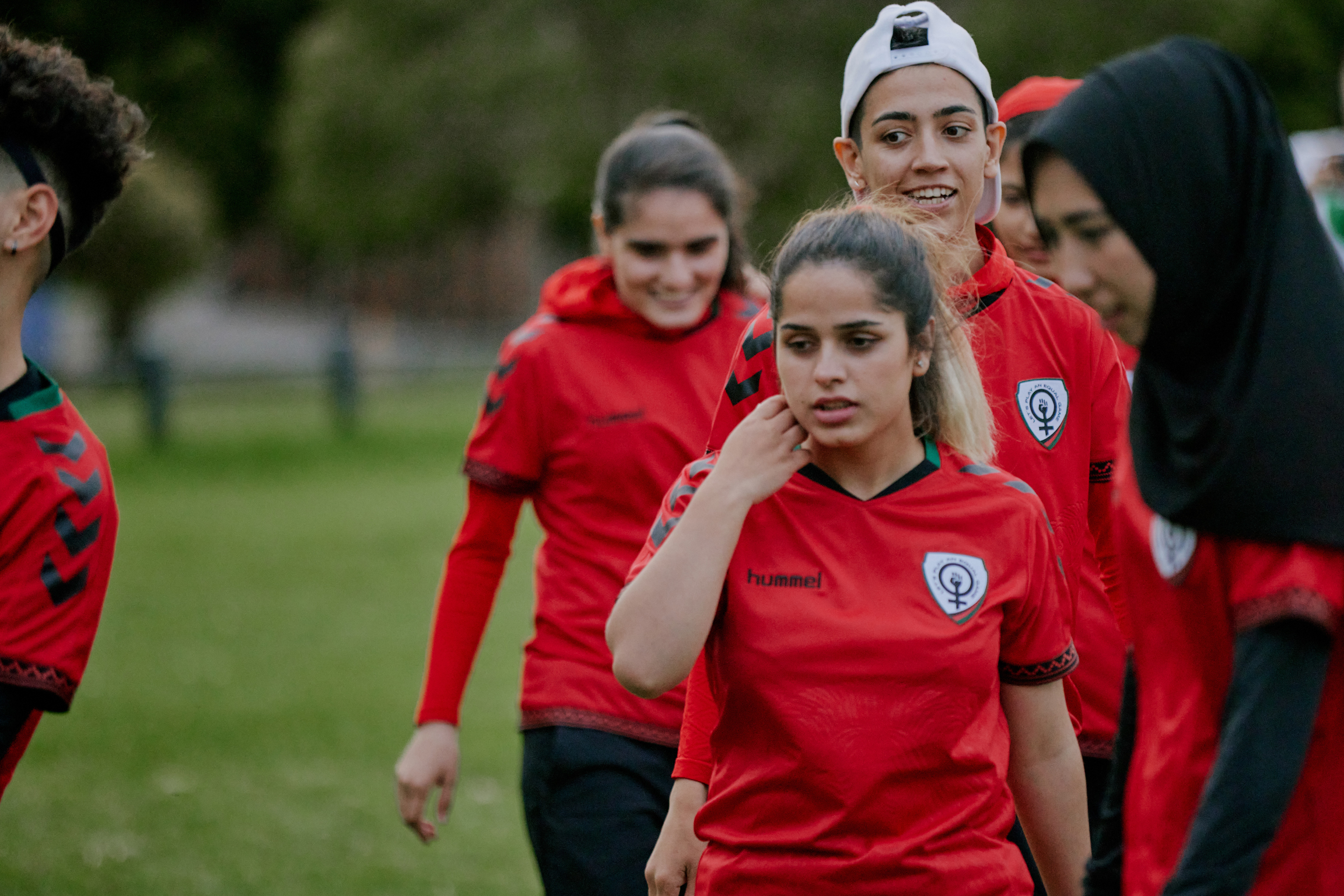 Group of young female footballers in red training kit with club badges walking together on grass pitch.