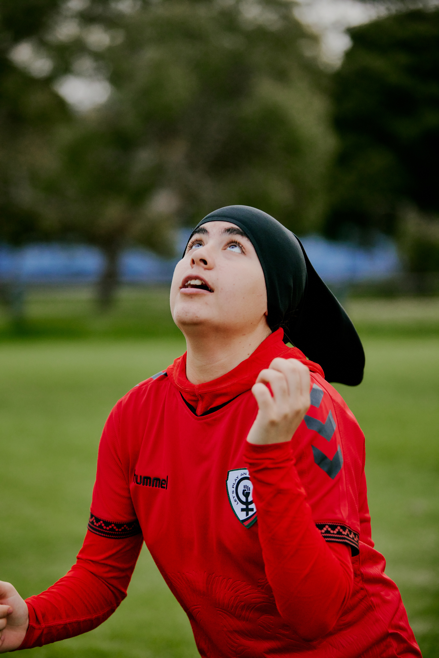 Woman in red football shirt and black hijab looking upwards on grass pitch with blurred trees in background.