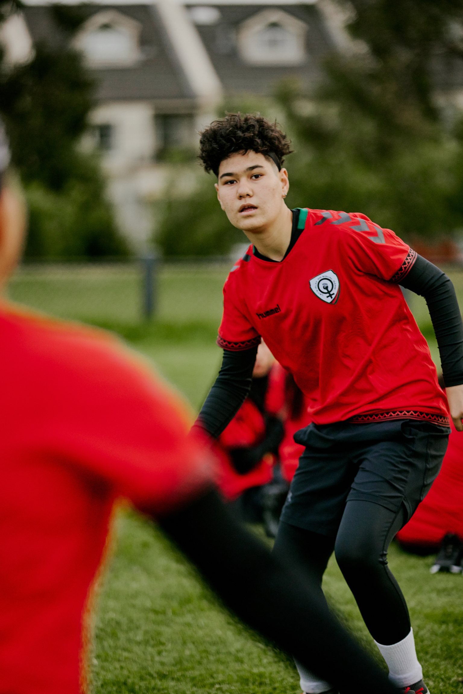 Young footballer in red and black kit during training, with teammates in foreground and residential buildings in blurred background.