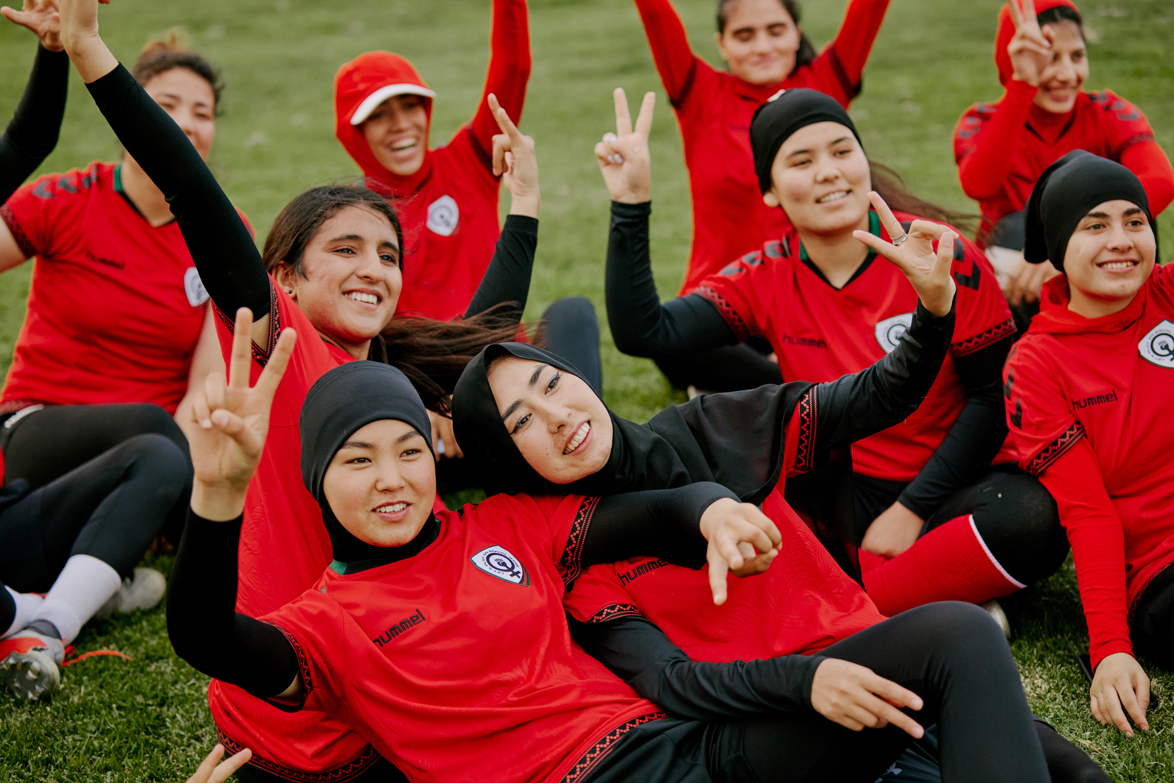 Group of female athletes in red and black sports kit celebrating on grass, raising arms and making victory signs whilst smiling.