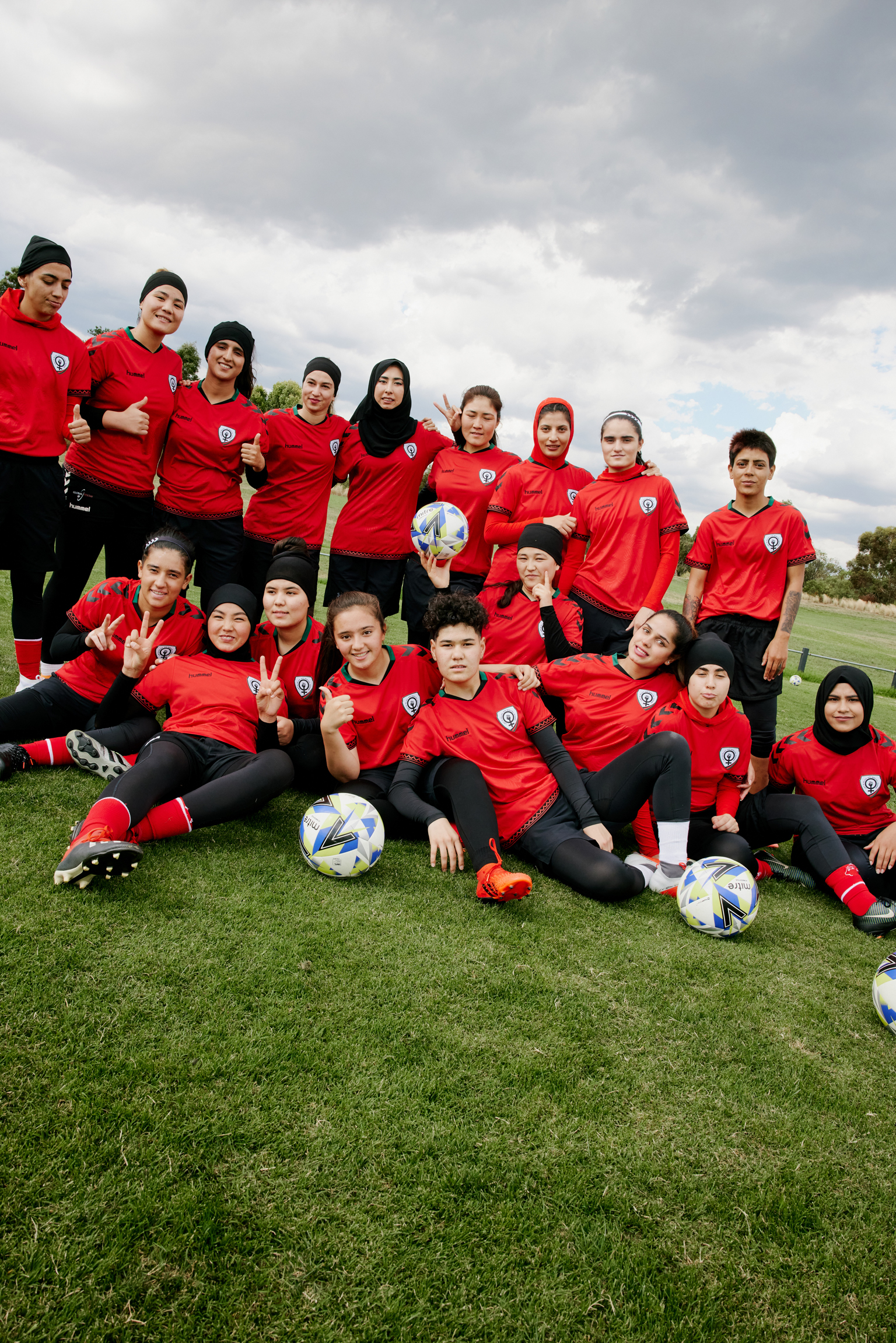 Youth football team in red training tops and black bottoms posing on grass pitch with footballs under cloudy sky.