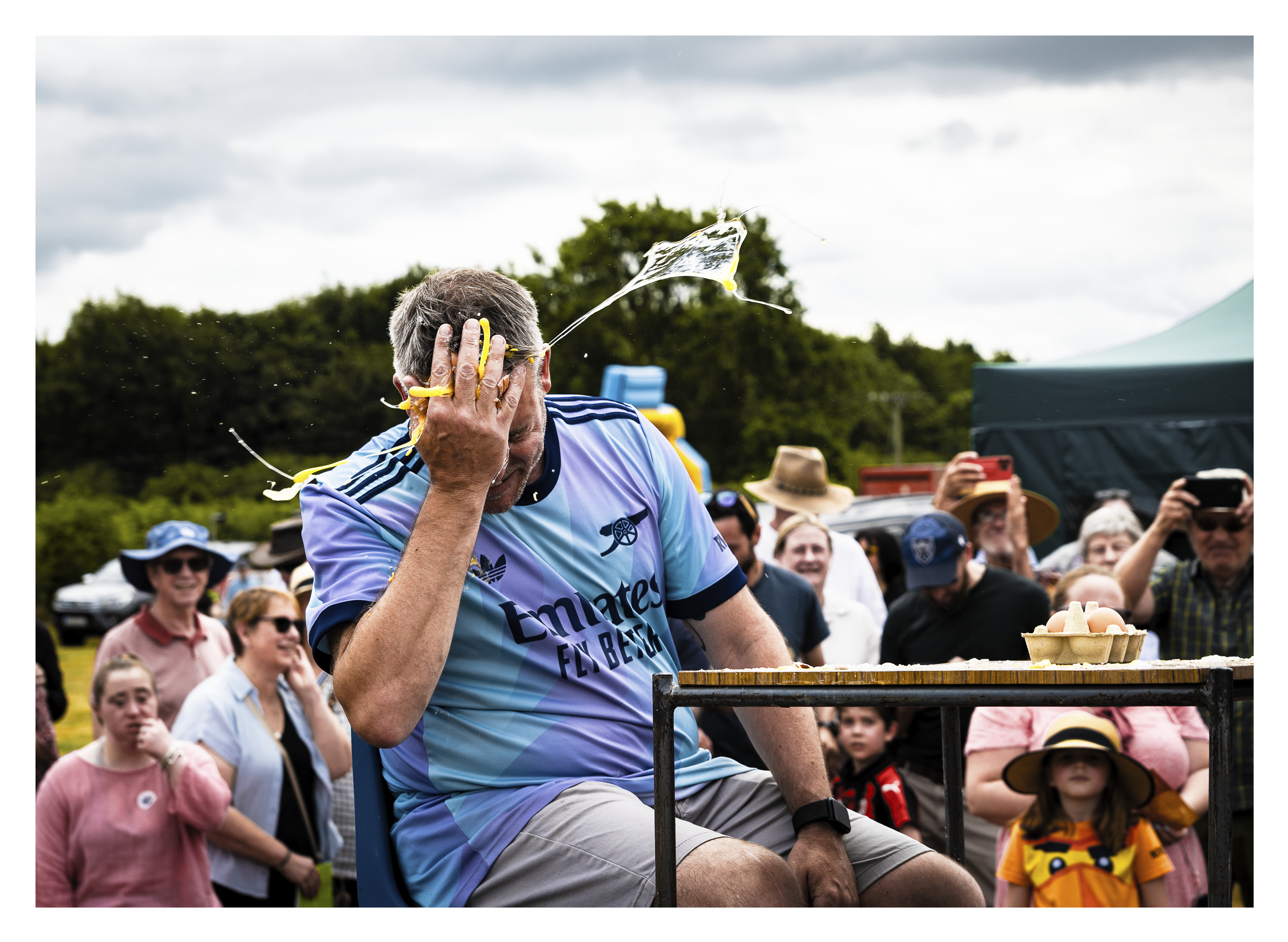 Man in light blue sports shirt raises hand to face whilst seated, with crowd of spectators watching in background under cloudy sky.