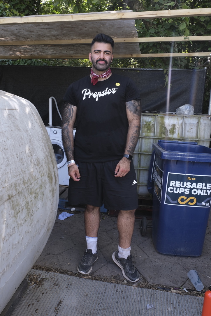 Bearded man with tattoos wearing black "Prepster" t-shirt and red bandana stands near recycling bins and wooden fence outdoors.