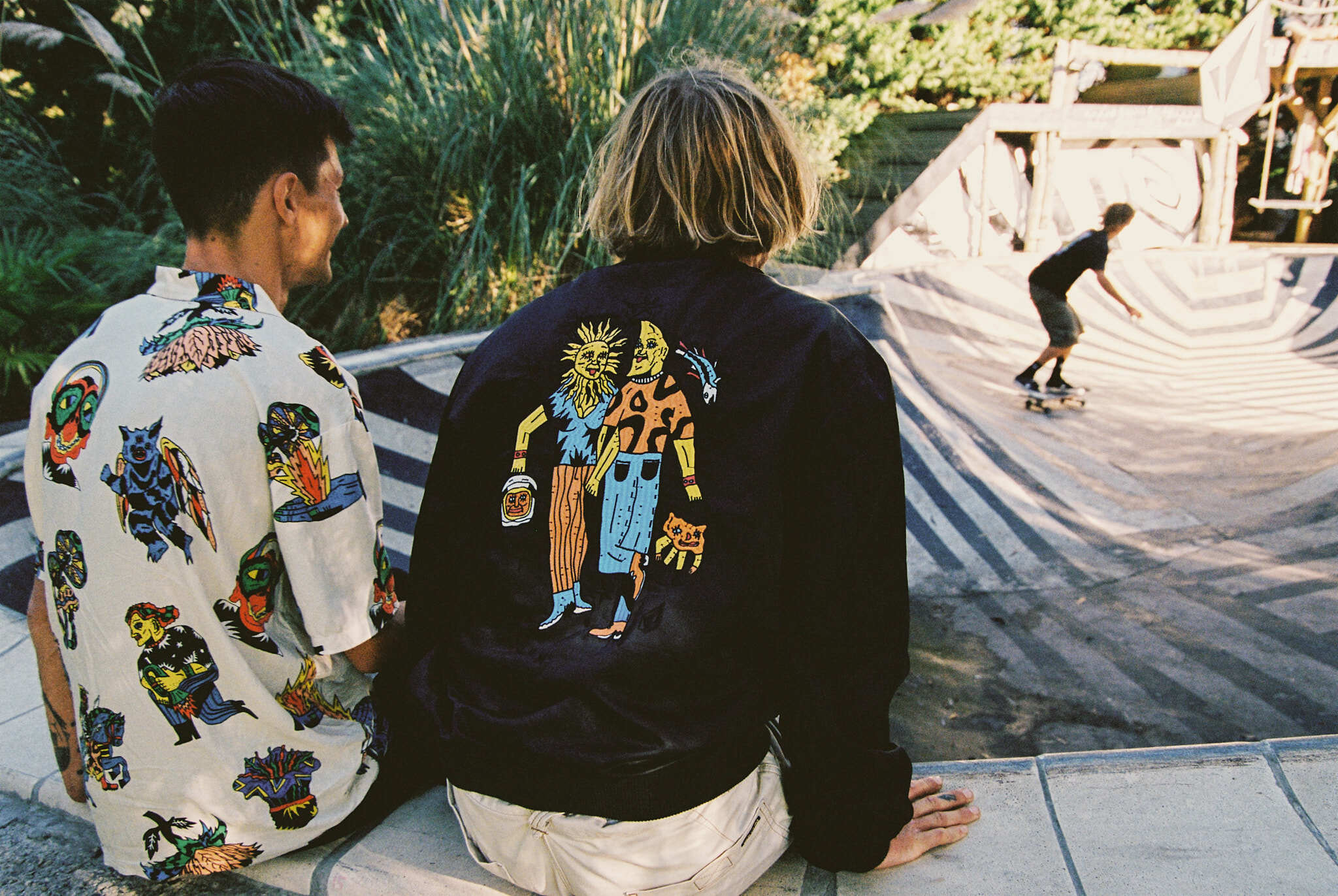 Colourful embroidered jackets worn by two people, with skateboarder visible in background. Bright colours and graphic designs on the clothing.
