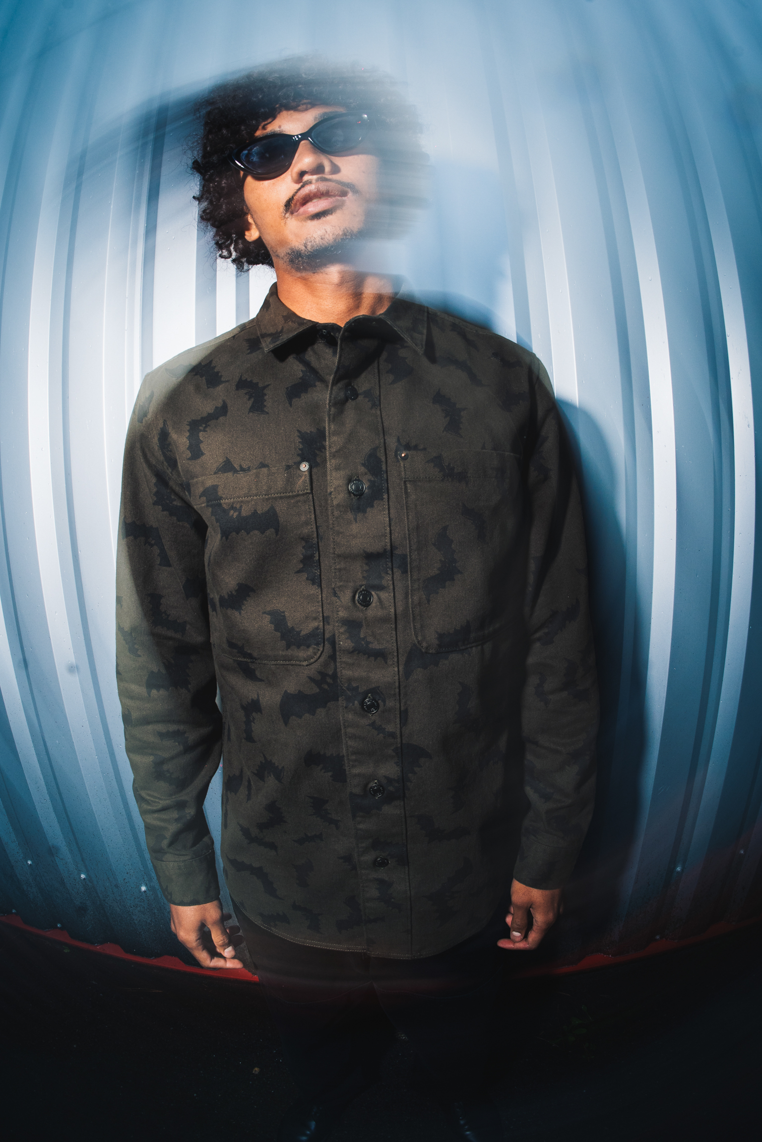 Man with curly hair wearing dark sunglasses and olive green camouflage shirt, standing in blue-lit curved tunnel or corridor.