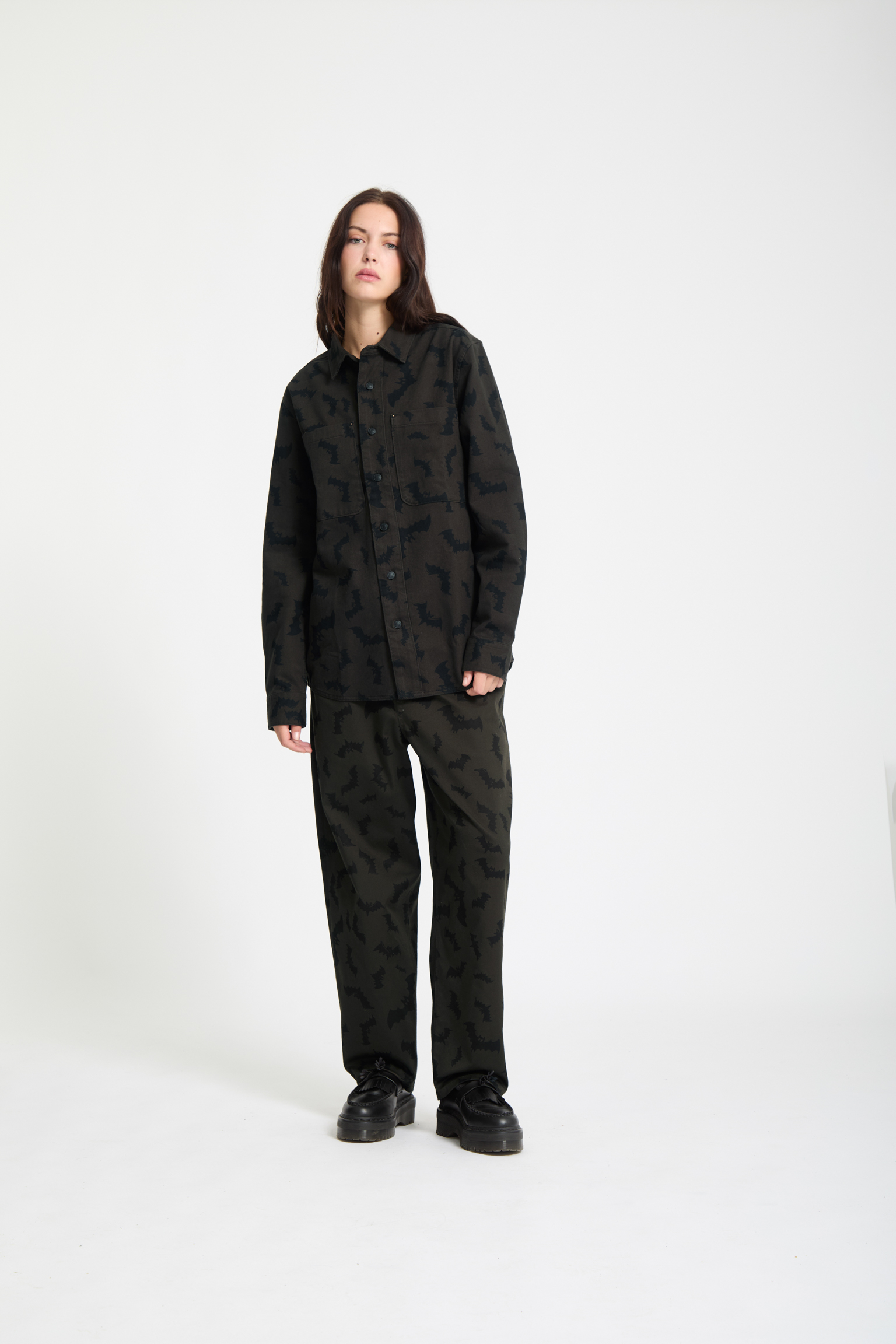 Woman wearing dark camouflage patterned shirt and trousers with black trainers, standing against light grey background.