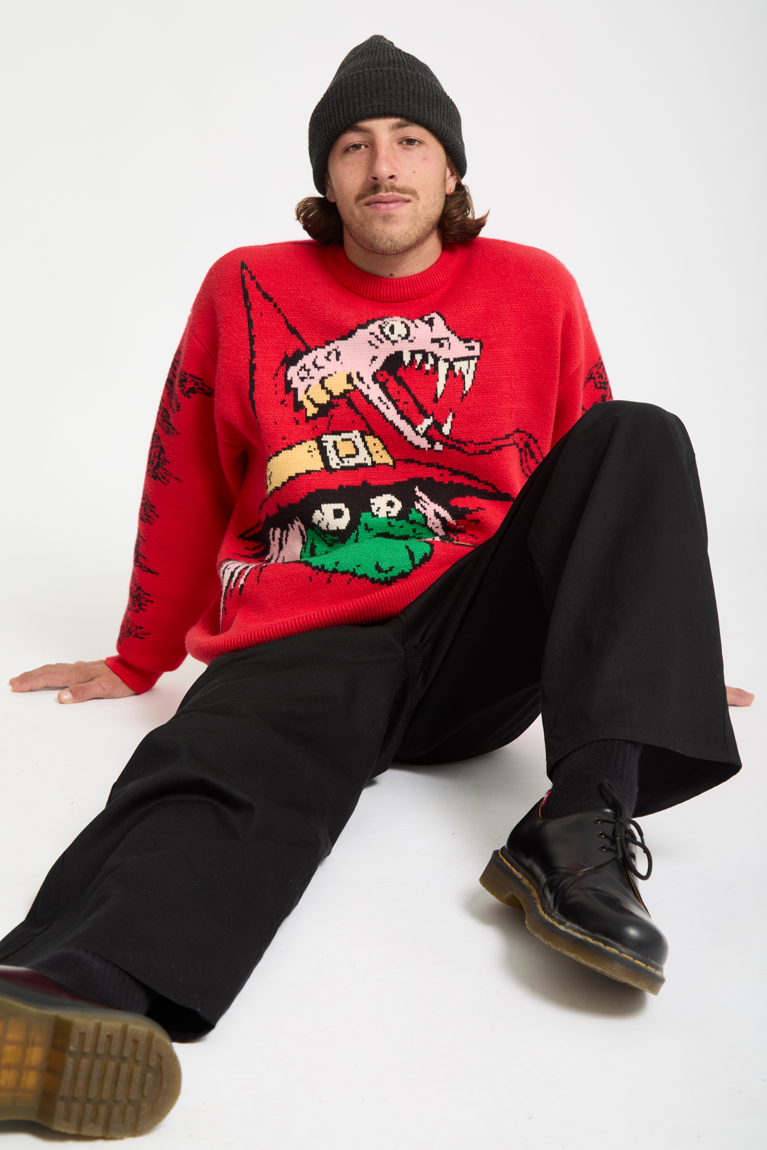 Man in black beanie and red Christmas jumper with dinosaur design, wearing black trousers and boots, sitting on white background.