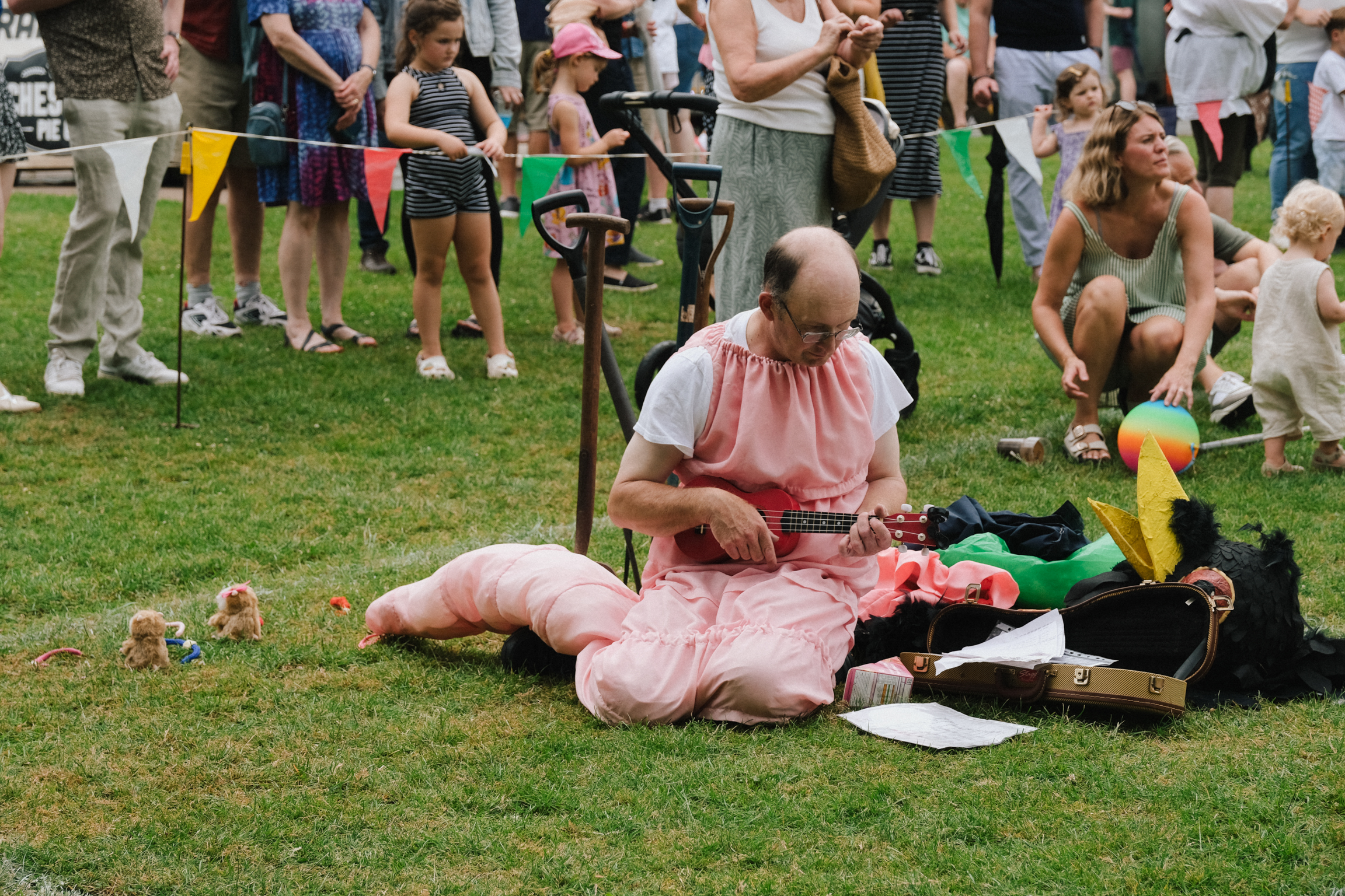 Man in pink tutu sitting on grass with pink pig prop, crowd with colourful bunting in background at outdoor event.