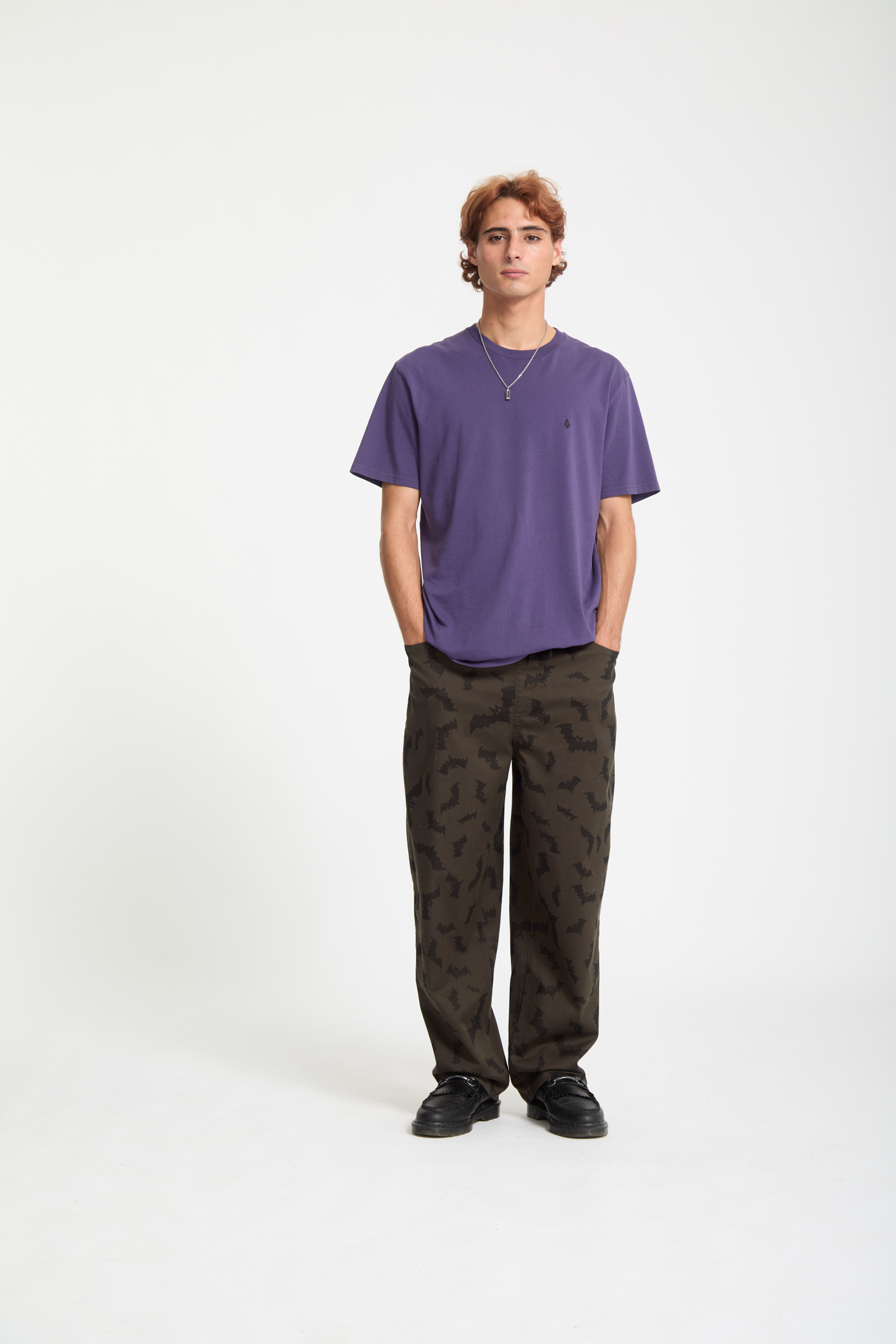 Young man with brown curly hair wearing purple t-shirt, dark camouflage trousers and black trainers against white background.