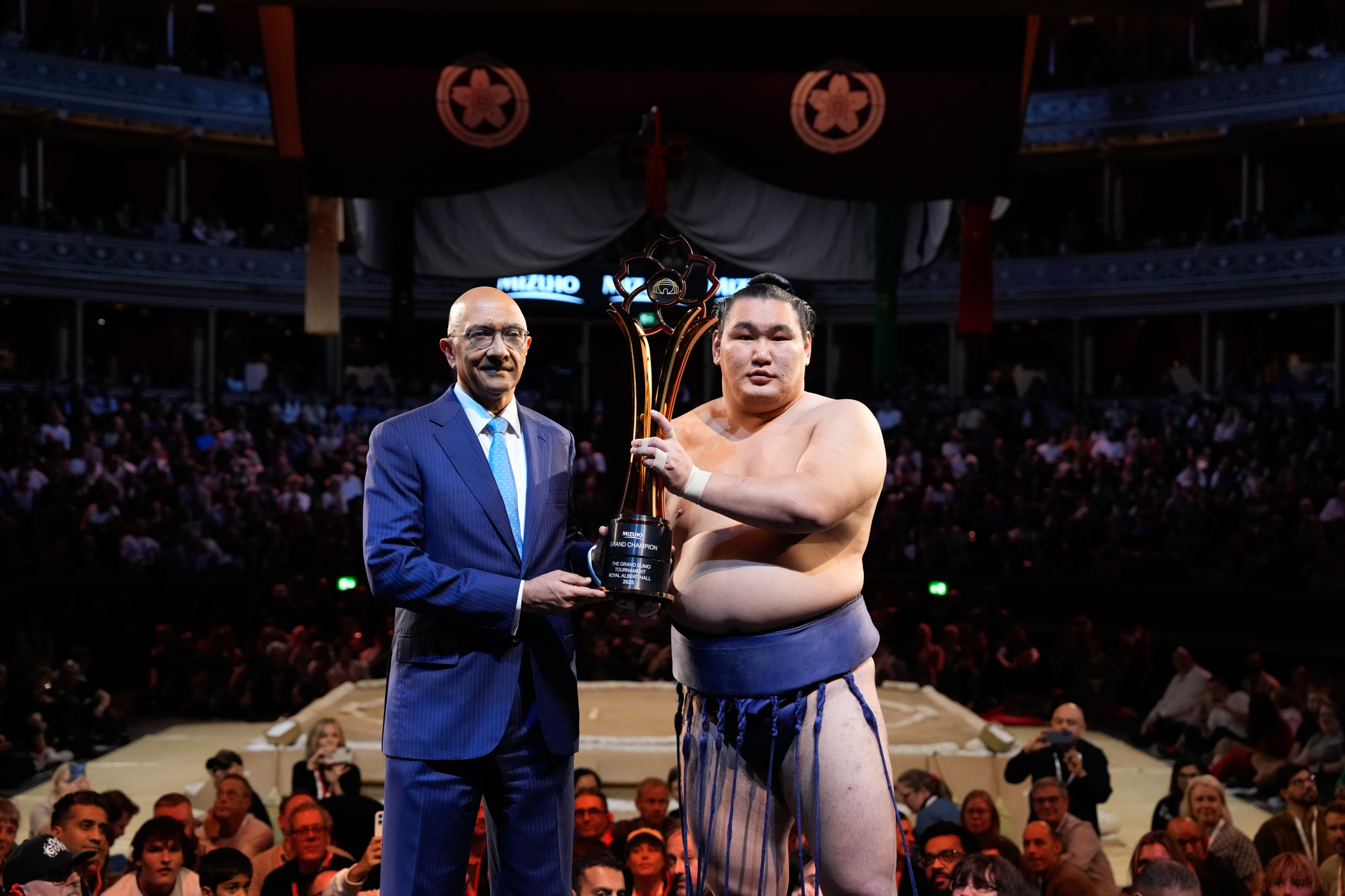 Man in blue suit presenting trophy to shirtless wrestler in blue shorts on sumo ring, crowd watching from tiered seating.